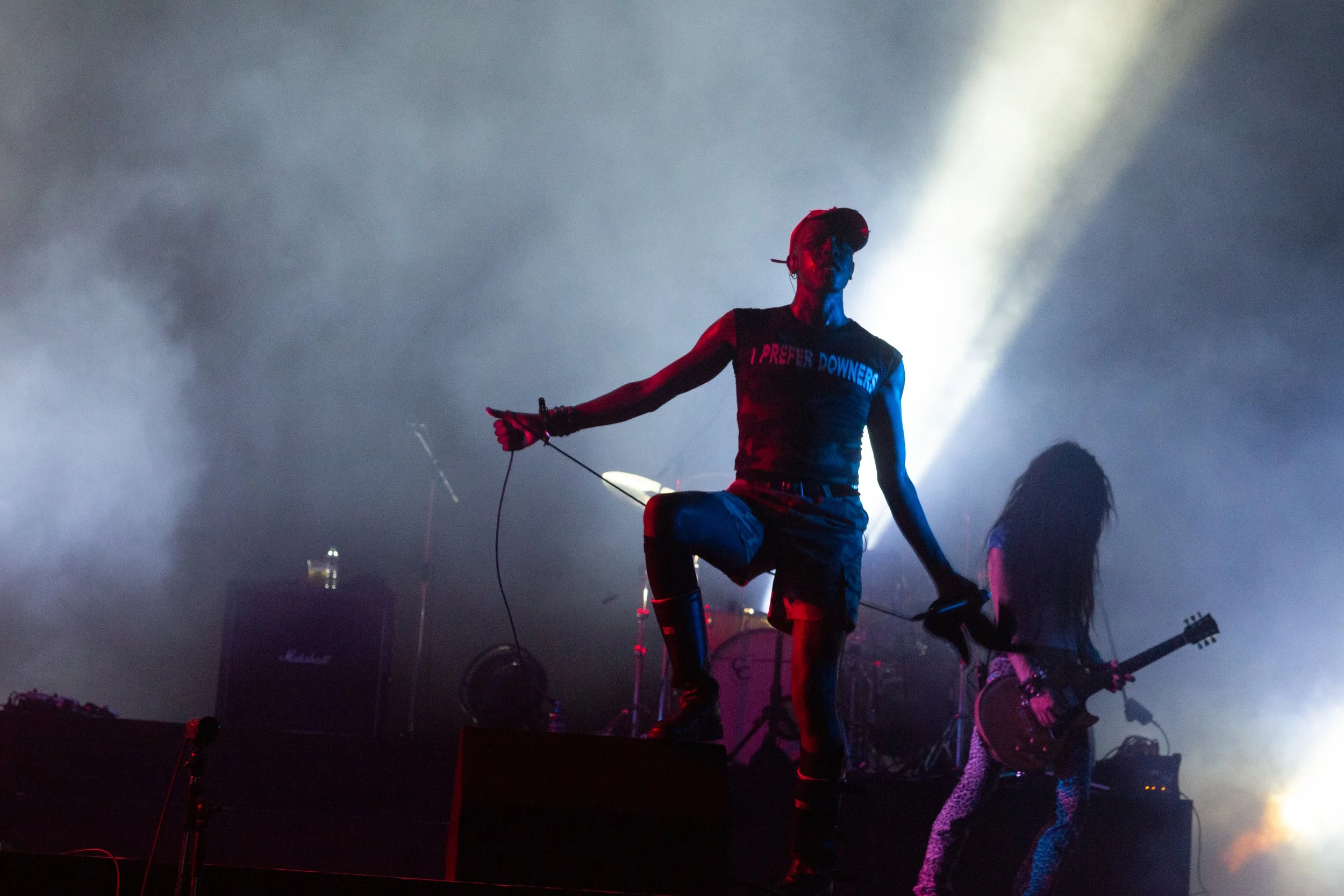 Musician on stage holding microphone and leaning on one leg with band members and equipment in background, illuminated by dramatic lighting.