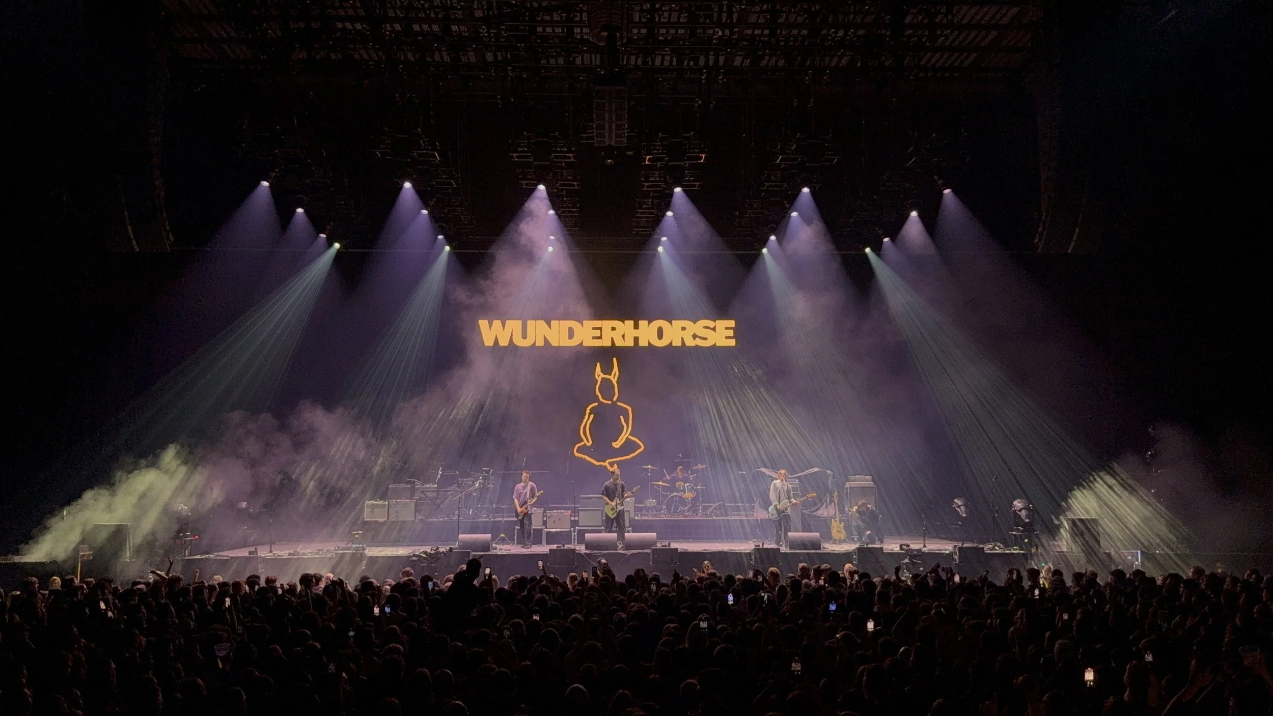 A live concert with a large crowd, featuring band members on stage with the sign 'WUNDERHORSE' and a dog silhouette logo behind them, illuminated by purple and white stage lights.