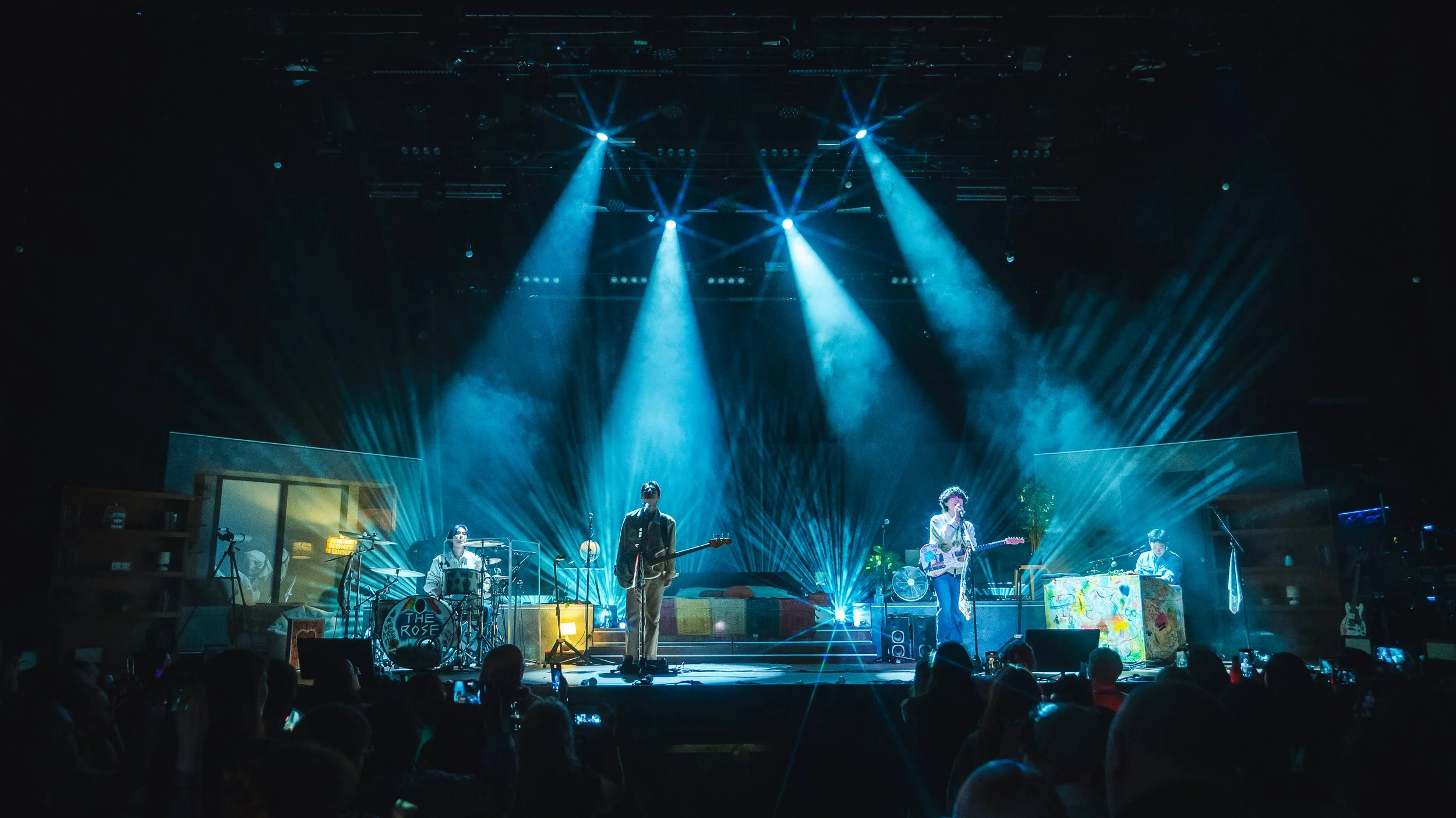 A band performs on a stage illuminated with blue and white spotlights, with audience members watching in the dark.