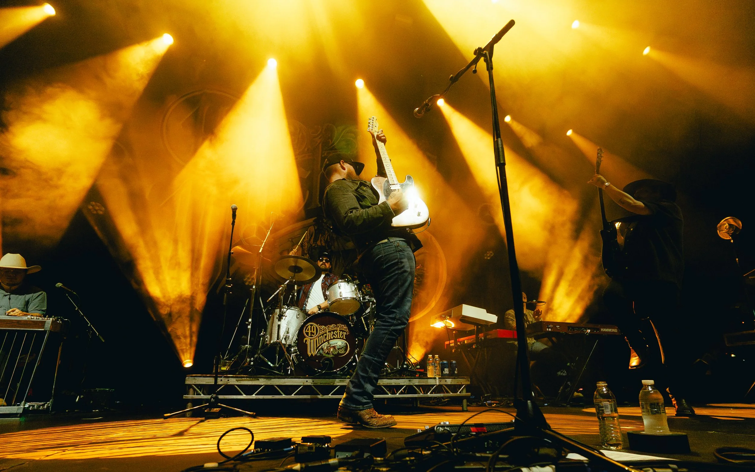 Musicians performing on stage with yellow and orange lighting, including a guitarist in the center, a drummer behind, and another guitarist to the right, with water bottles on the floor.
