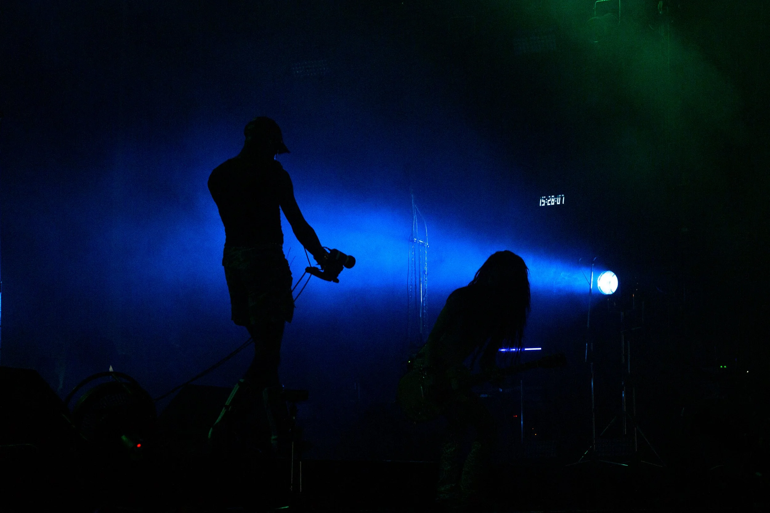 Silhouettes of two musicians, one standing and one crouching, on a stage with blue and green lighting, holding instruments, during a performance.