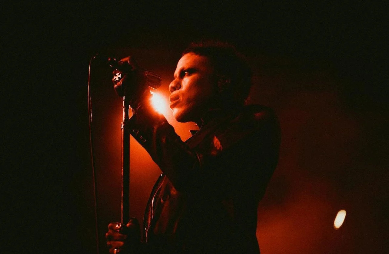 A man with short hair and a leather jacket singing into a microphone on stage with red and orange backlighting.