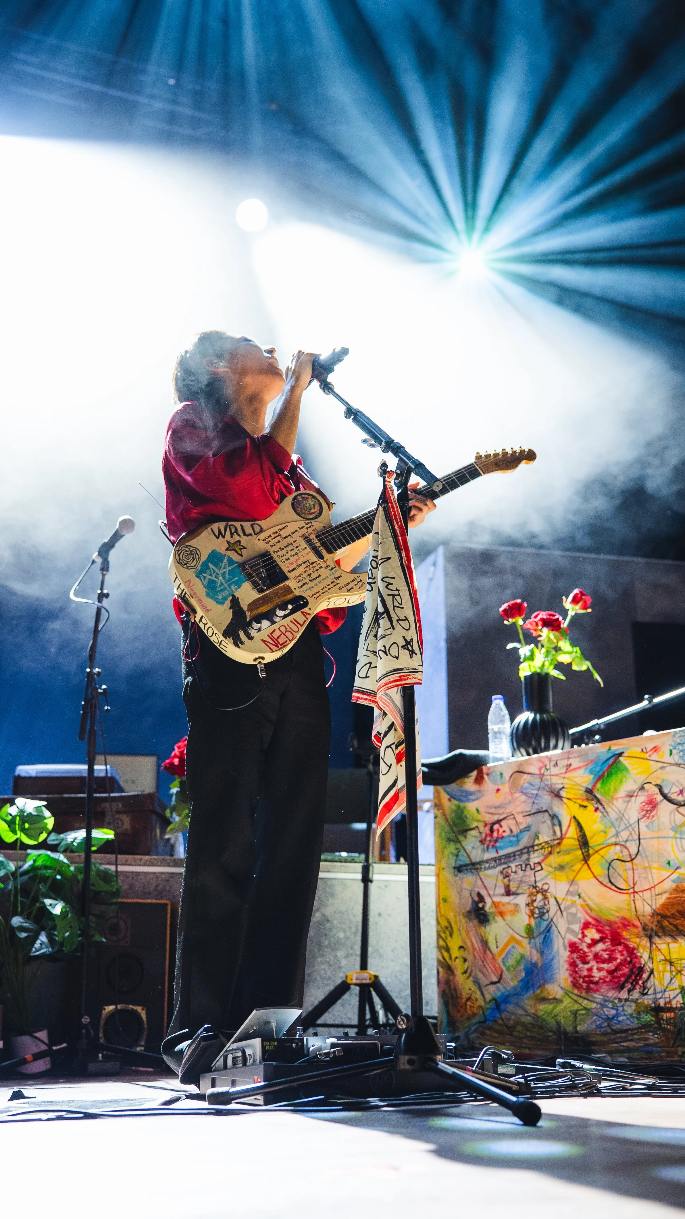 A man singing and playing an electric guitar on stage with bright lights and fog effects, decorated with colorful artwork and flowers.