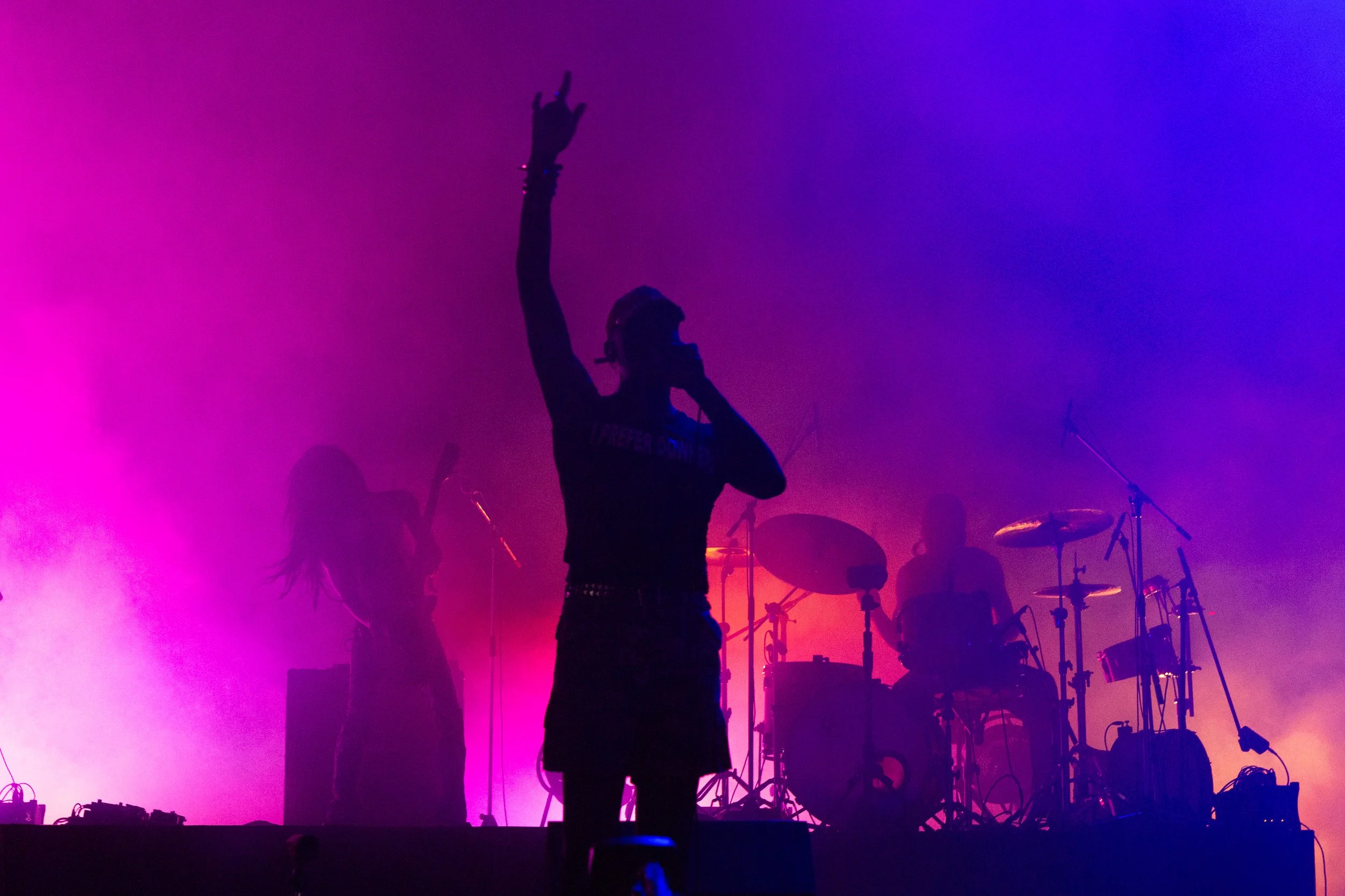 Silhouette of a singer on stage holding a microphone and gesturing with one hand, with a band playing in the background, illuminated by pink and purple stage lights.