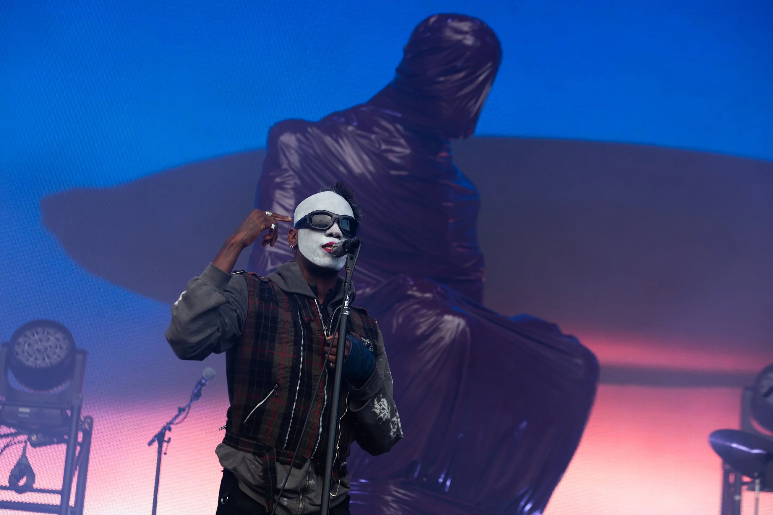 Performer with painted face, wearing sunglasses, headphones, and a plaid jacket, singing into a microphone on stage with a large statue of a seated figure in the background against a pink and blue sky backdrop.