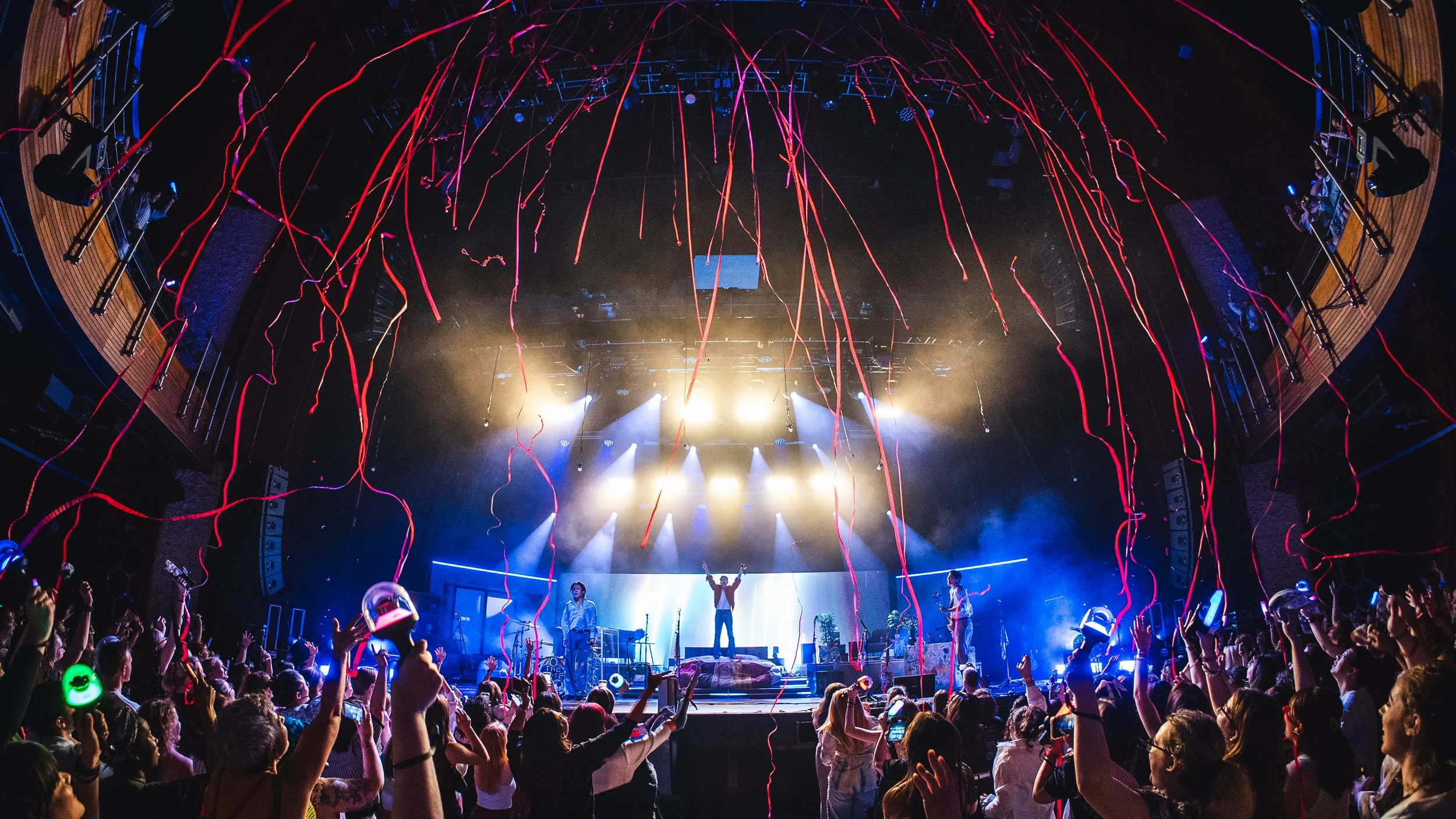 Concert stage with performers, colorful lights, and streamers, audience holding glow sticks, and a vibrant atmosphere.