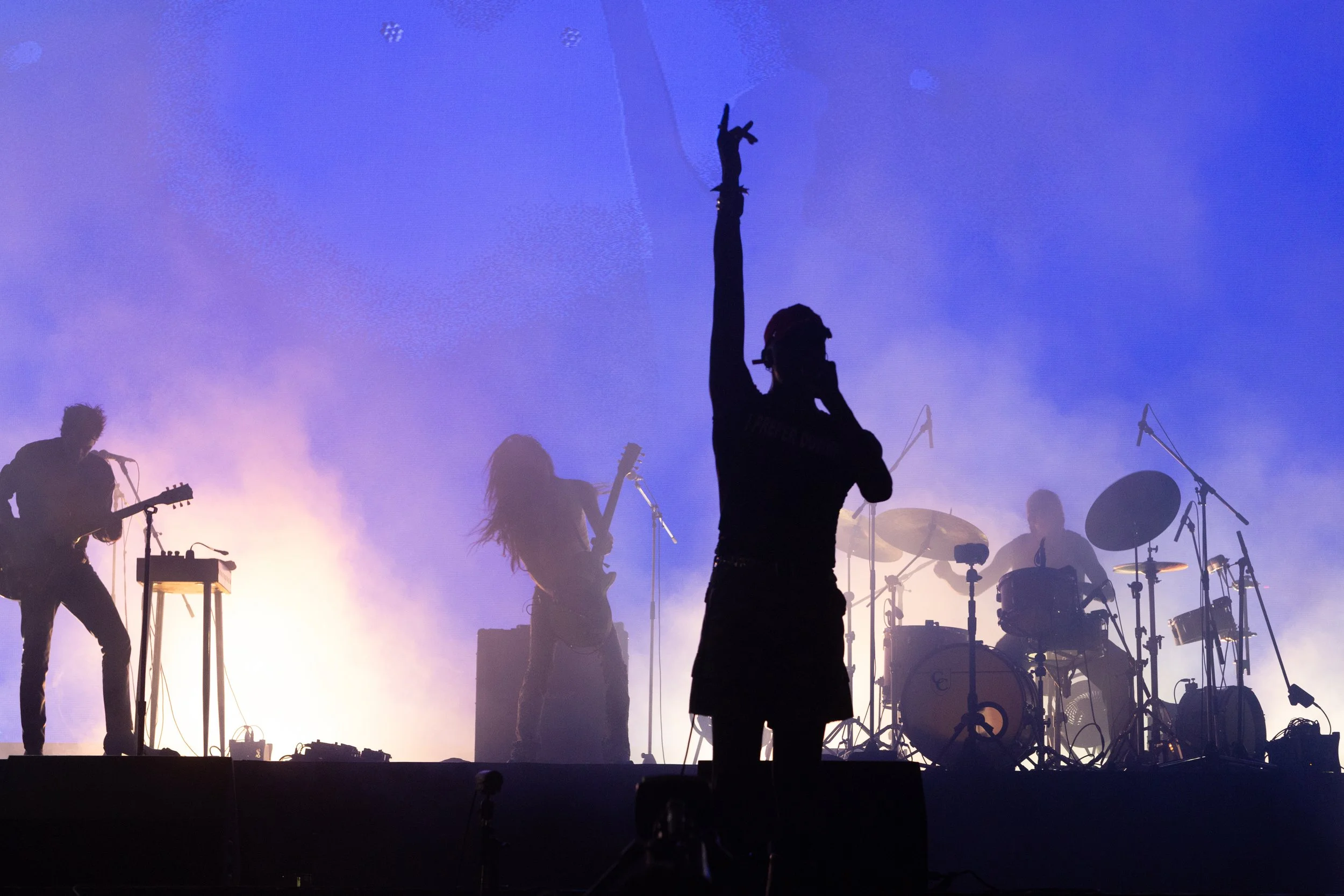 Silhouette of a band performing on stage with purple and yellow lights, includes a singer with one arm raised, a guitarist, a drummer, and a keyboardist, with musical equipment and microphones.
