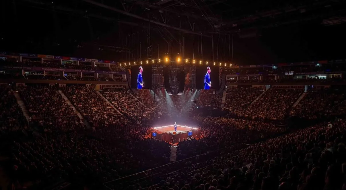 A large indoor arena filled with spectators, with a stage in the center illuminated by bright lights. A presenter is on stage speaking, with two large screens showing the presenter above the stage.