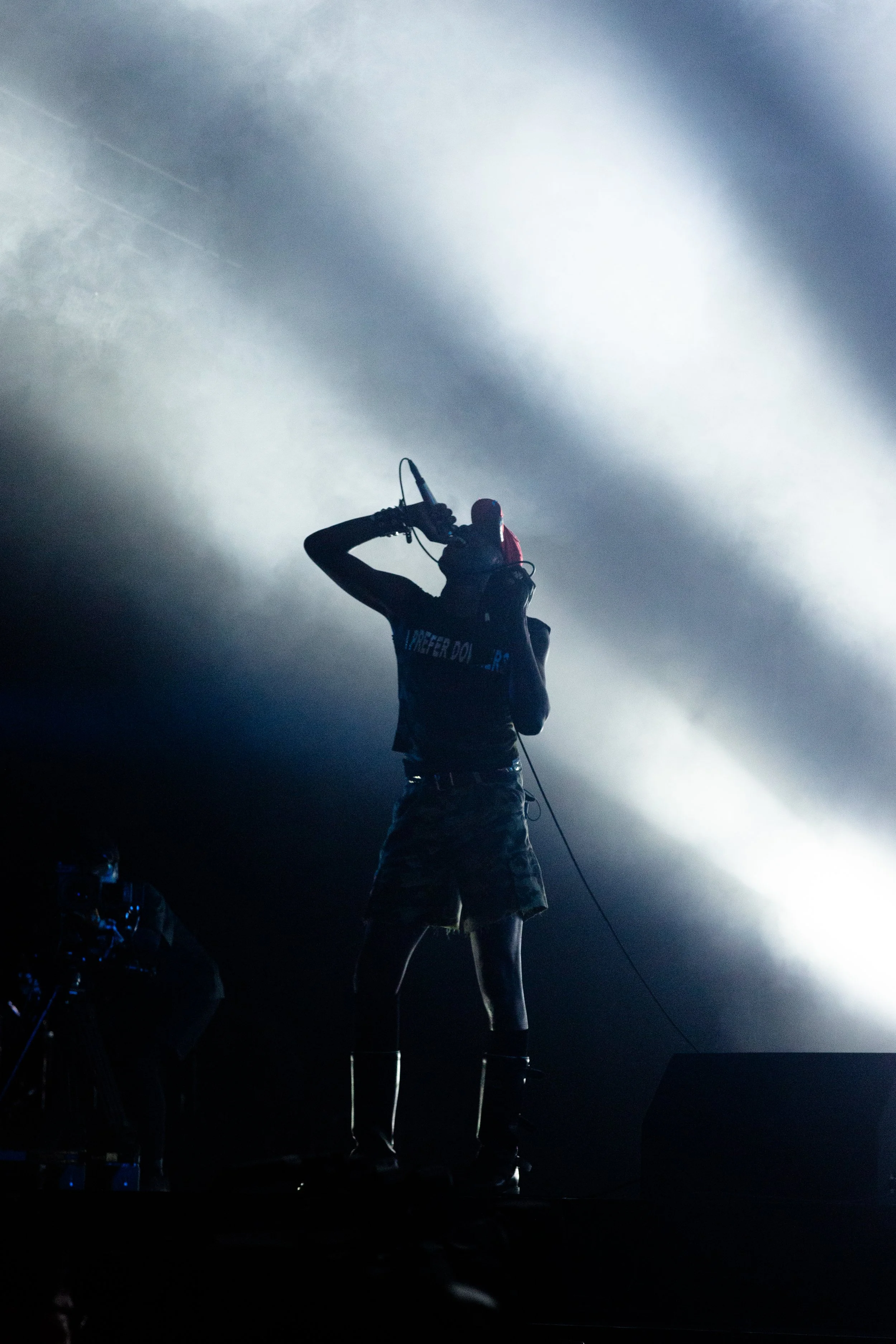 A performer singing on stage with a microphone, backlit by dramatic stage lighting, wearing camouflage shorts and long boots.