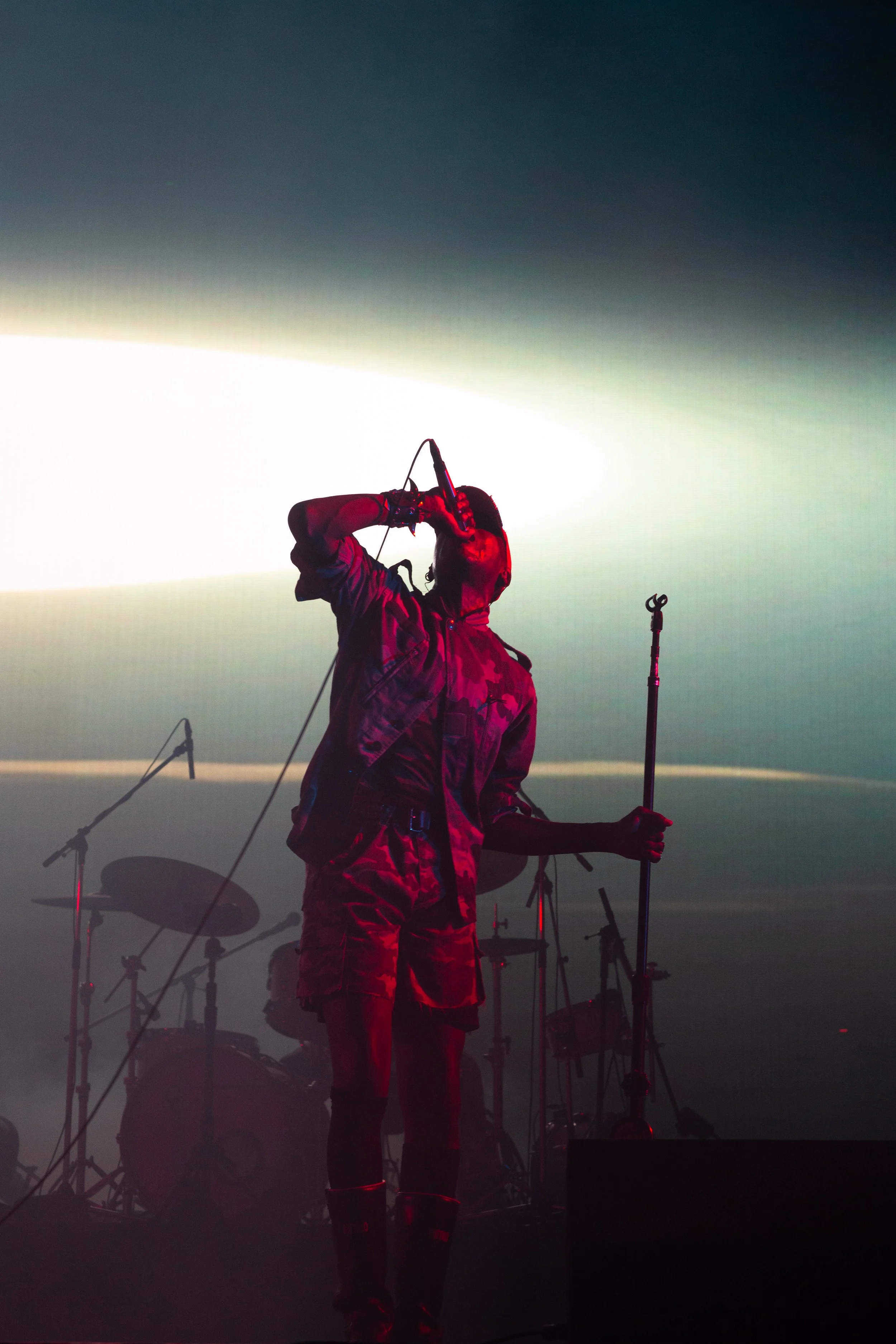 A musician performing on stage with stage lights, holding a microphone, and accompanied by drums and other musical equipment in the background.
