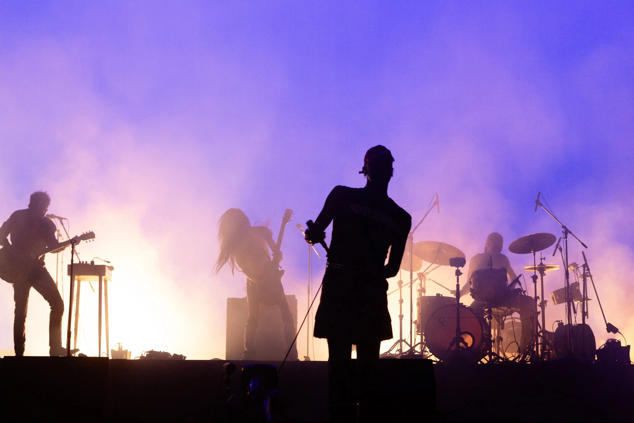 Silhouettes of a band performing on stage with purple and yellow lighting, including a guitarist, drummer, and singer, amidst stage fog.