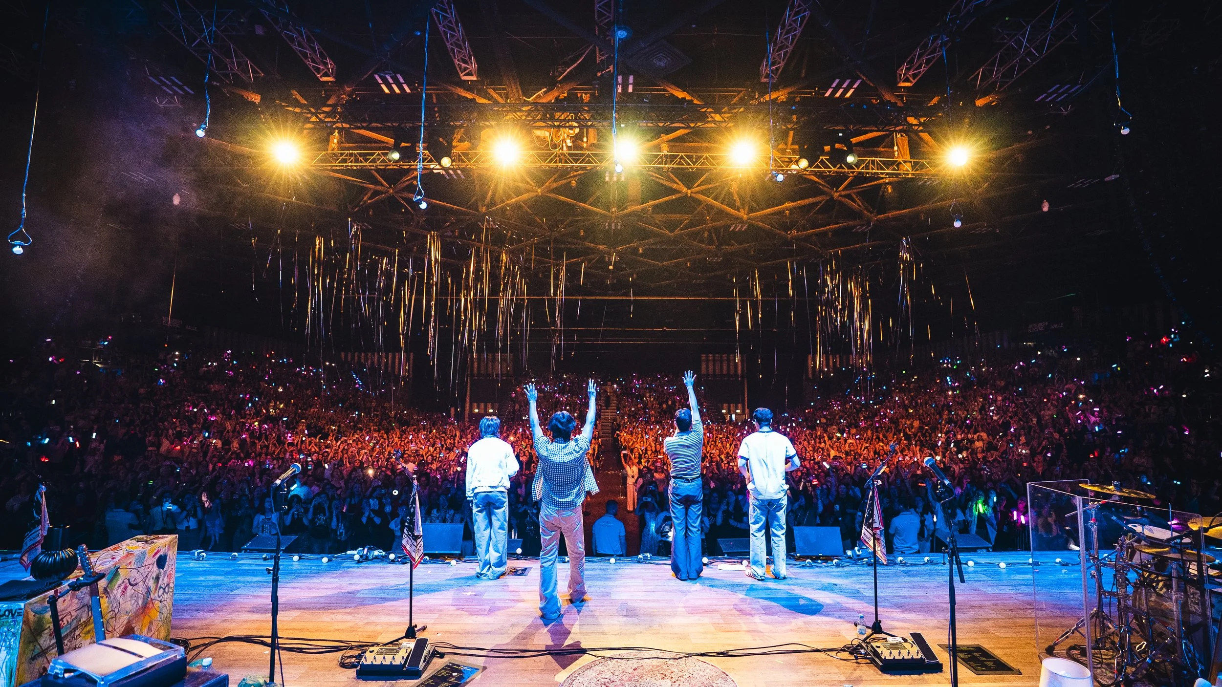 View from the stage of a concert with four performers facing a large audience, colorful lights, and decorative streamers hanging from the ceiling.