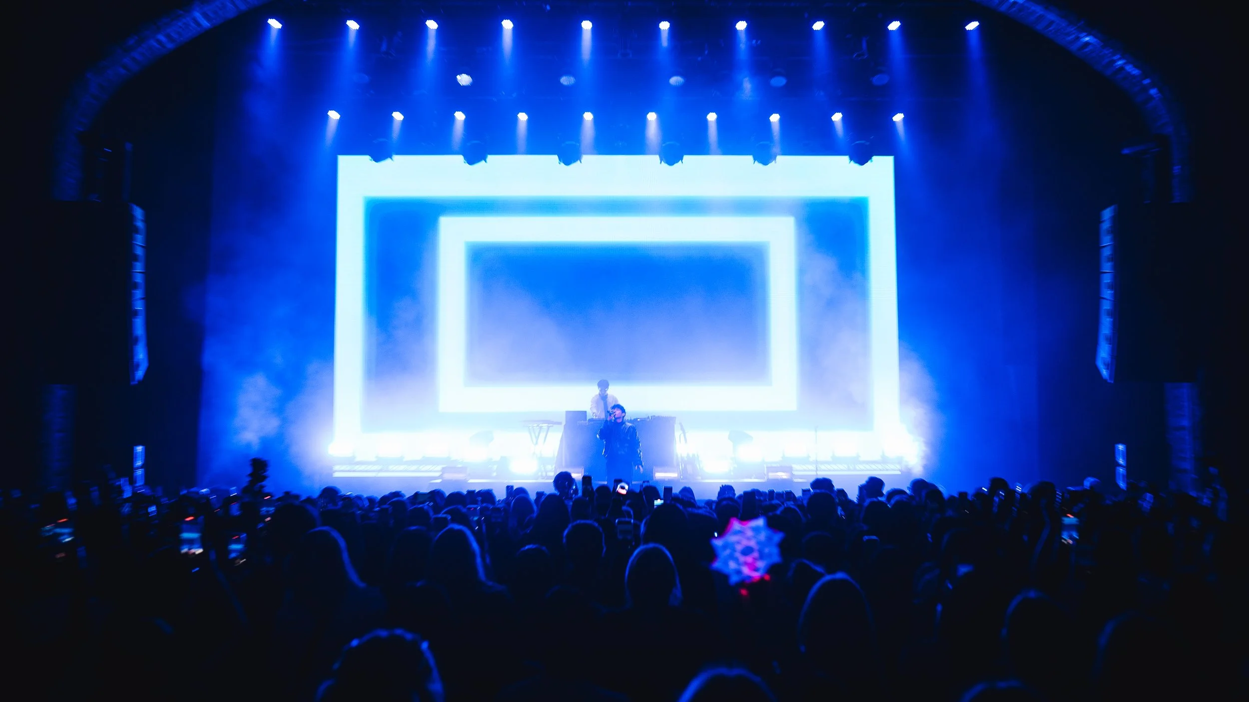 A concert stage illuminated with blue lights and large LED screens, with a crowd in the foreground.