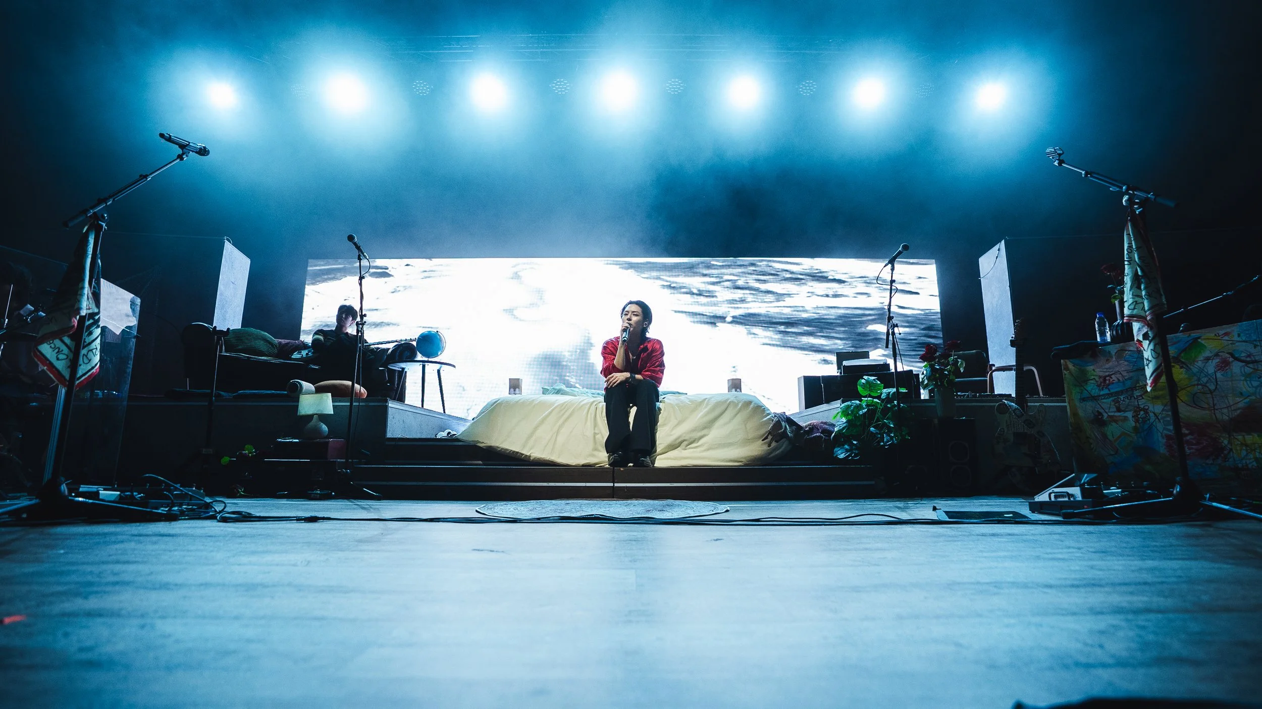 A man sitting on a bed on stage, holding a microphone, with artistic decor and equipment around her, and a large digital screen displaying abstract images in the background.