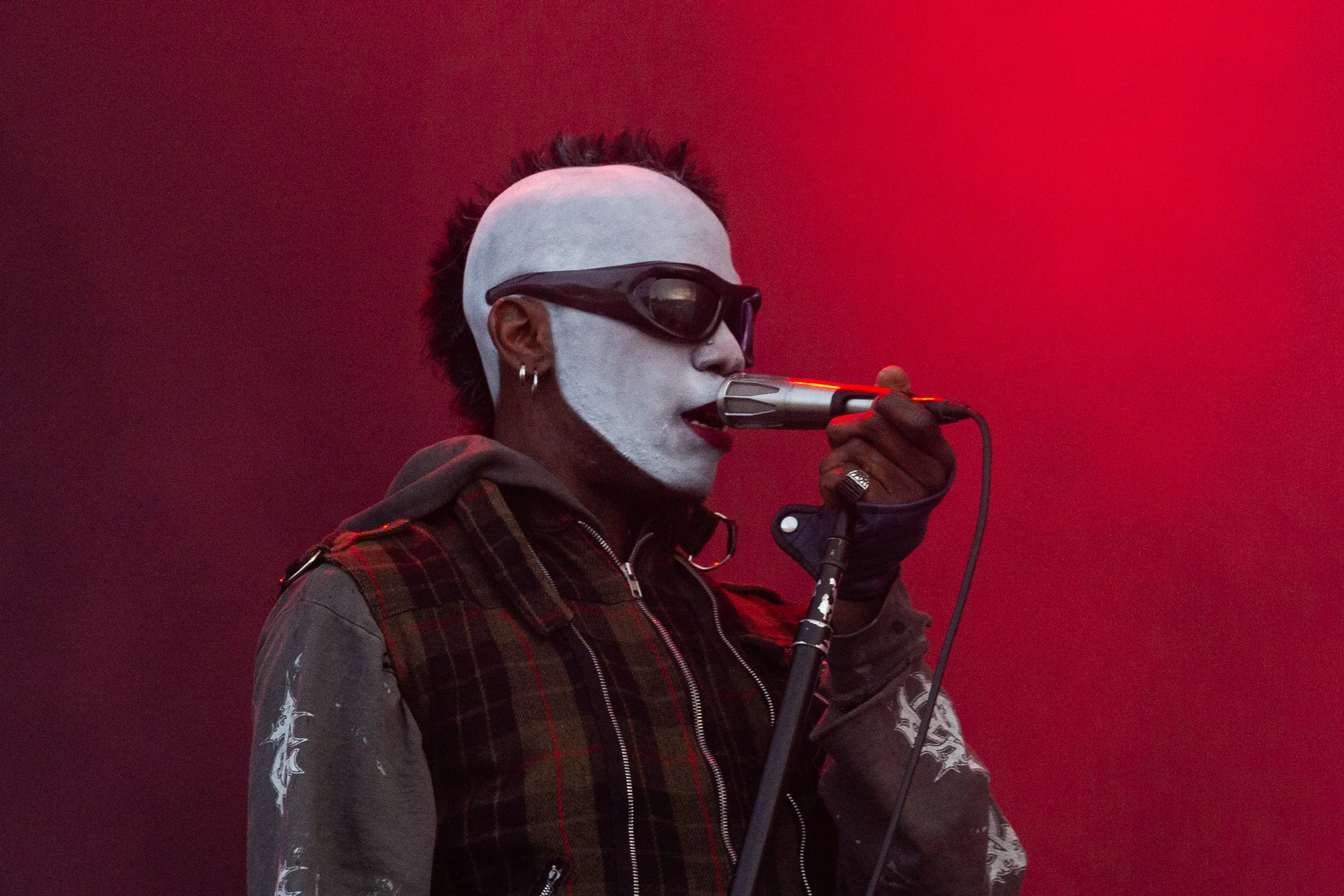A person with white face paint, black sunglasses, and earrings sings into a microphone on a red background.