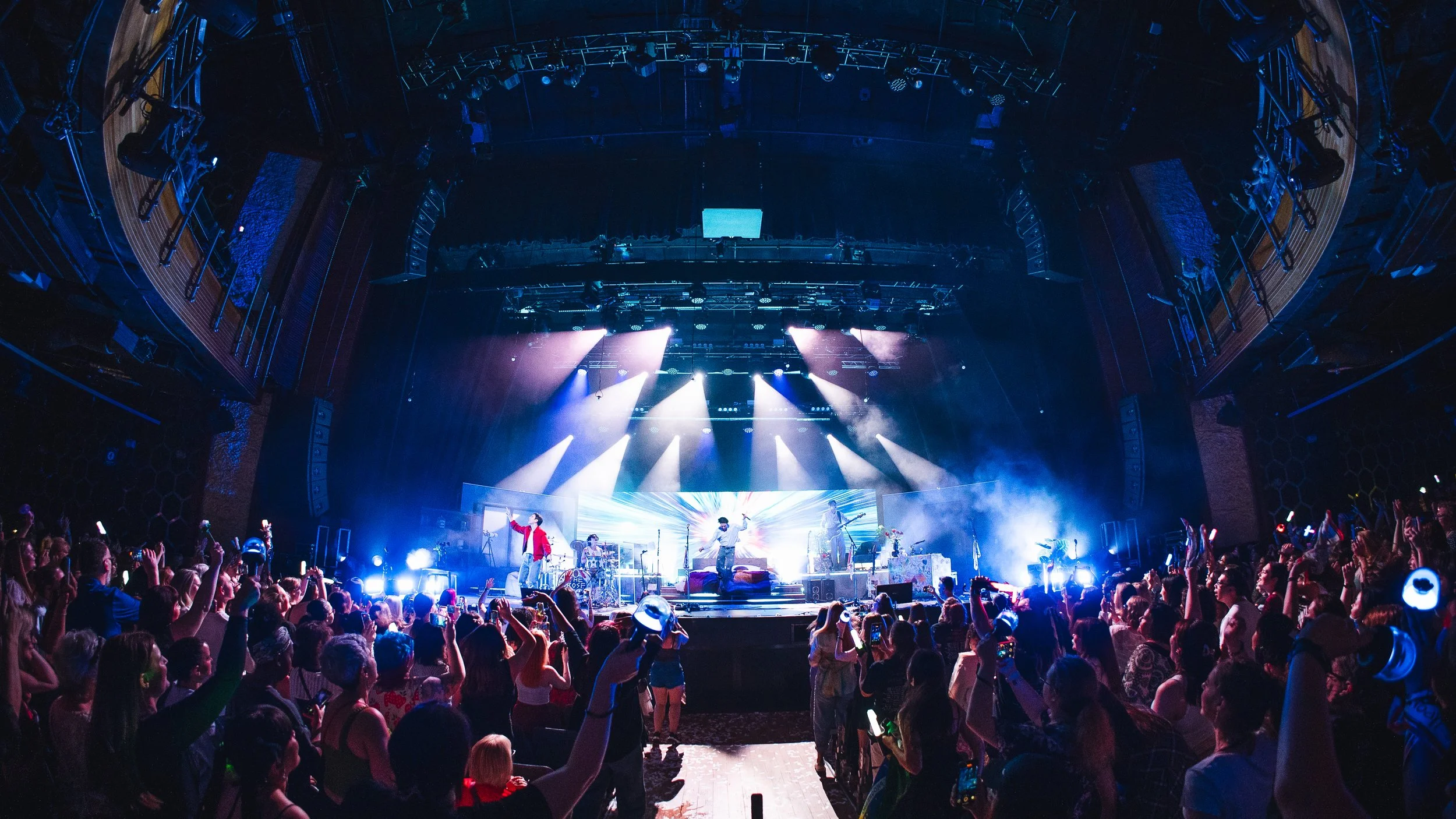 A lively concert with a band performing on stage and a large audience dancing and cheering, illuminated by colorful stage lights.