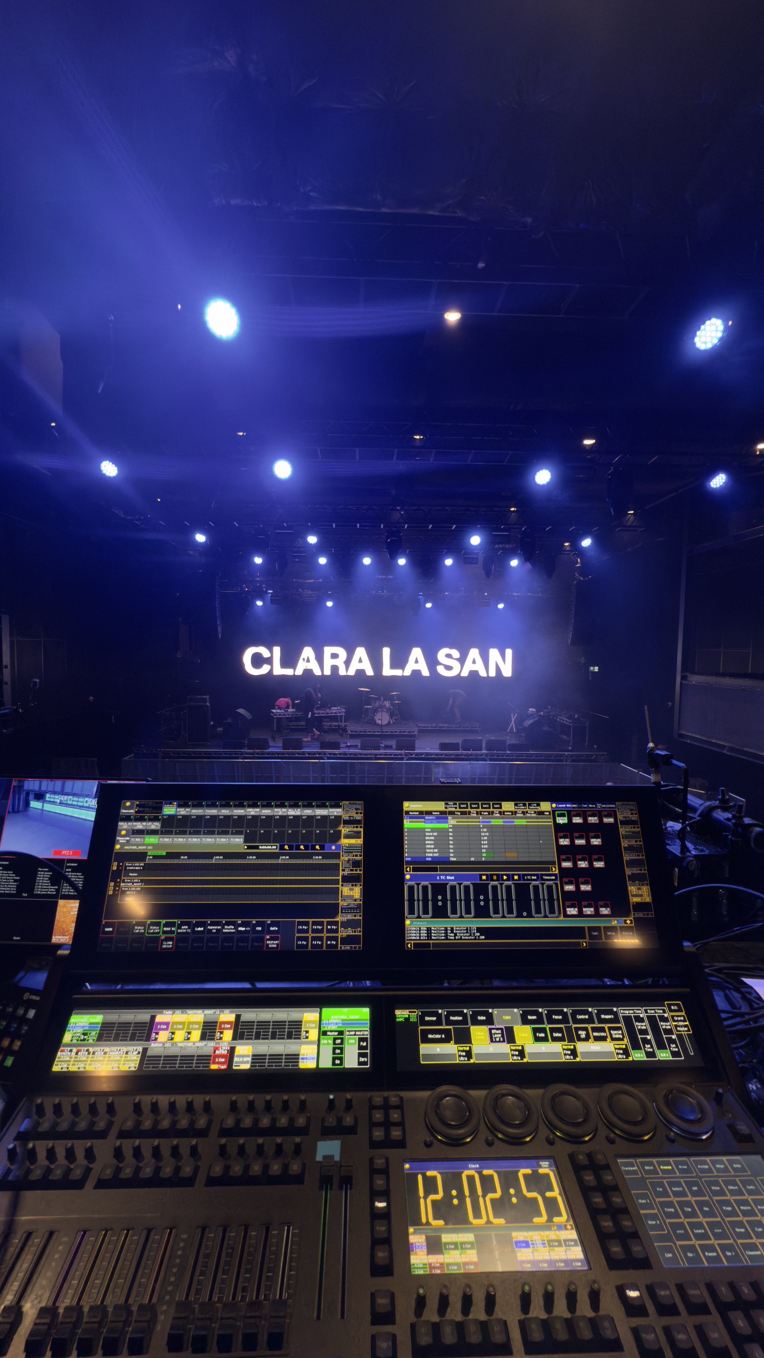 A view of a stage with a large LED screen displaying the name 'Clara La San' and a mixing console with various controls in the foreground, indicating a concert or event setup.