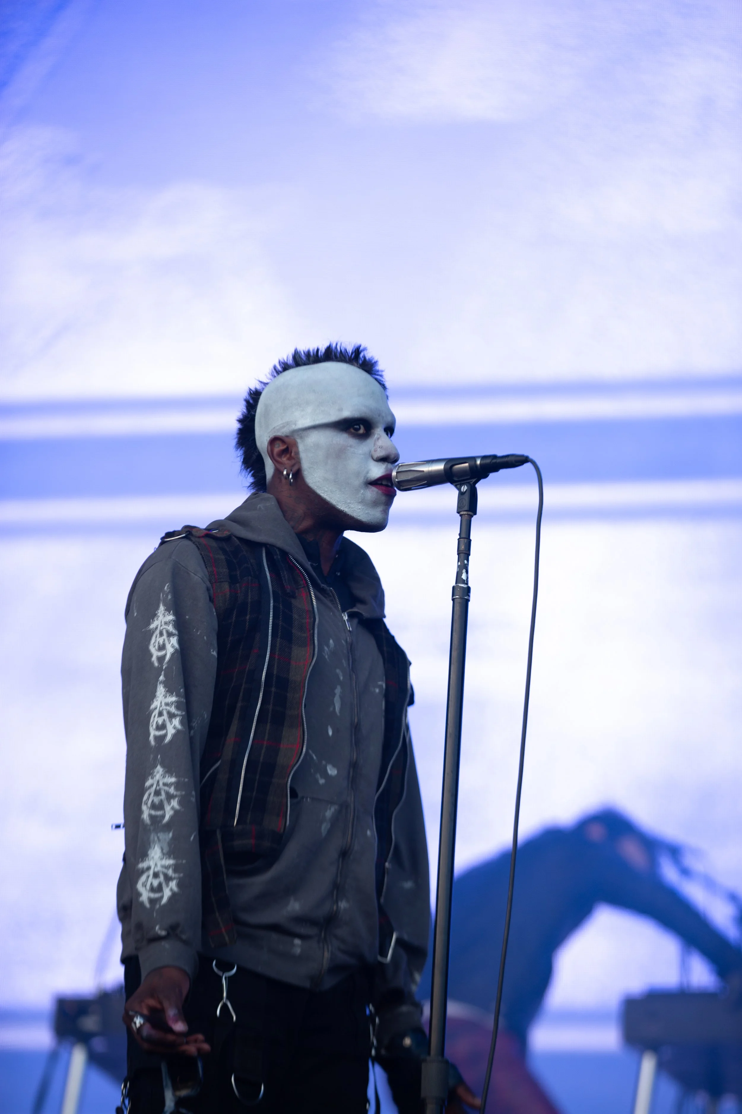 A man with white face paint, dark eye makeup, and a mohawk hairstyle singing into a microphone on stage.
