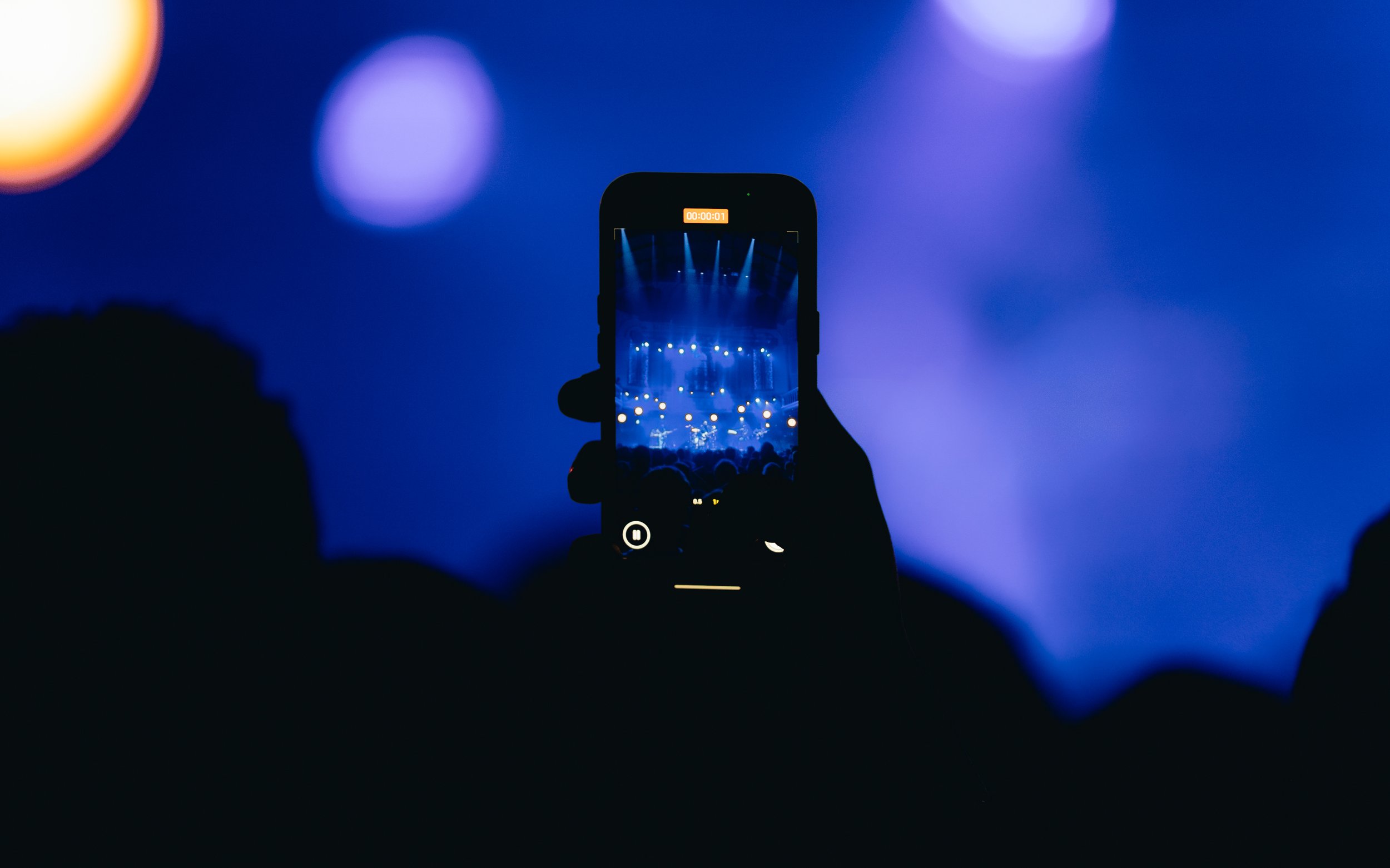 A person is holding a smartphone in front of a concert stage, capturing the performance which is illuminated with blue lights.