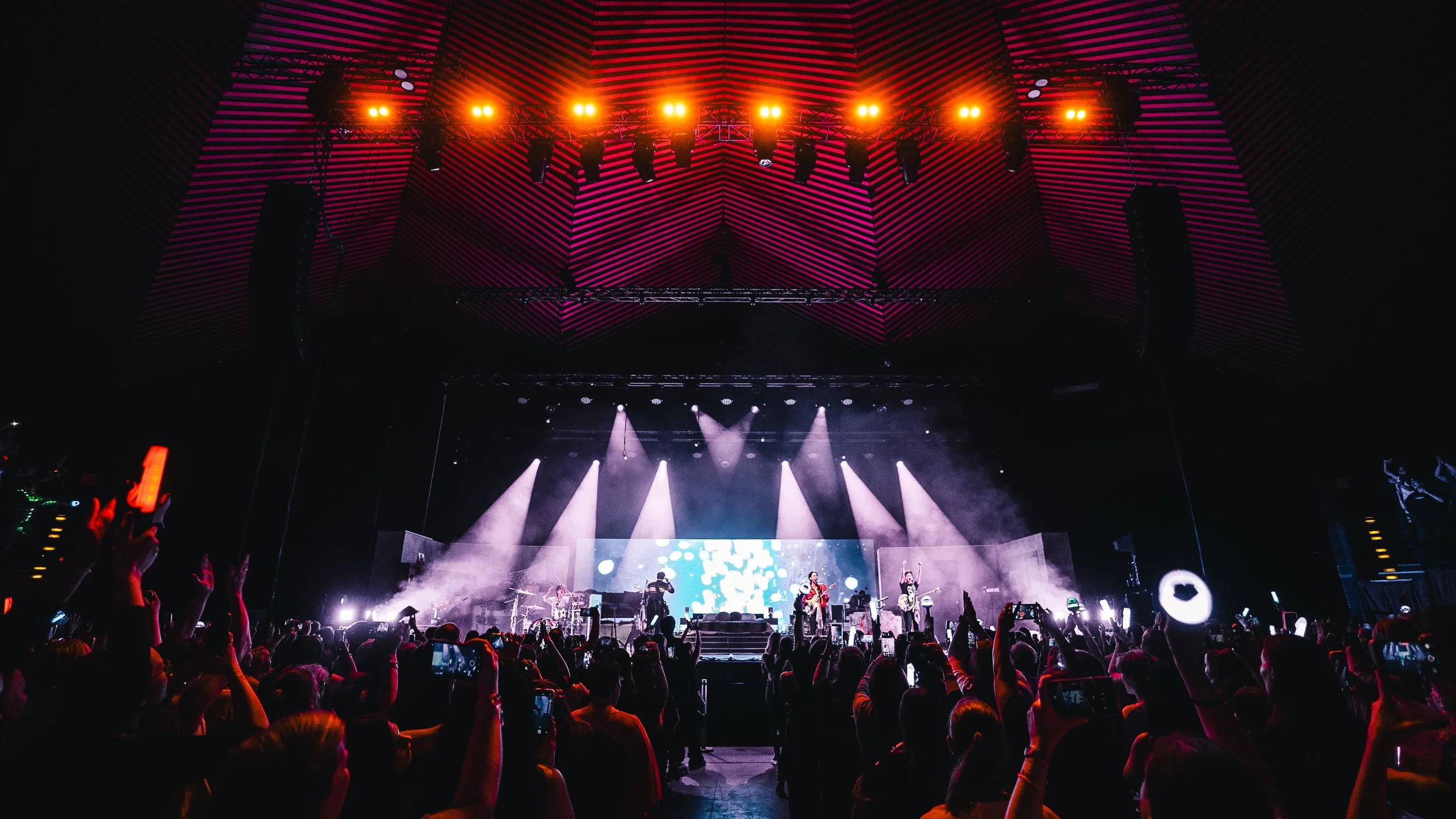 Concert stage with lights and digital screen, audience raising hands and taking photos at night.