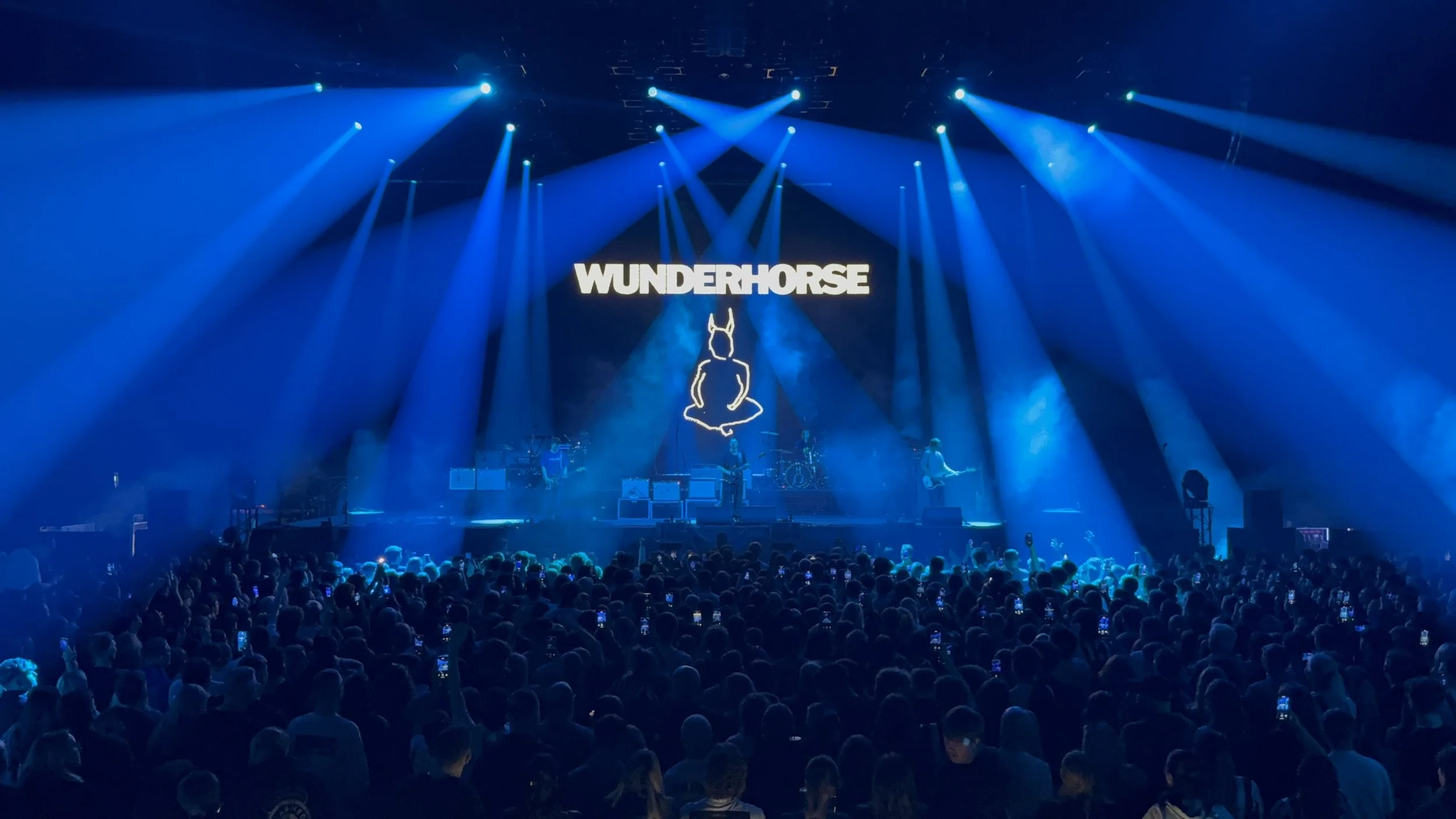 A concert stage with blue lighting, a large crowd in front, and a screen displaying the word 'WUNDERHORSE' above a line art image of a seated figure with animal ears.