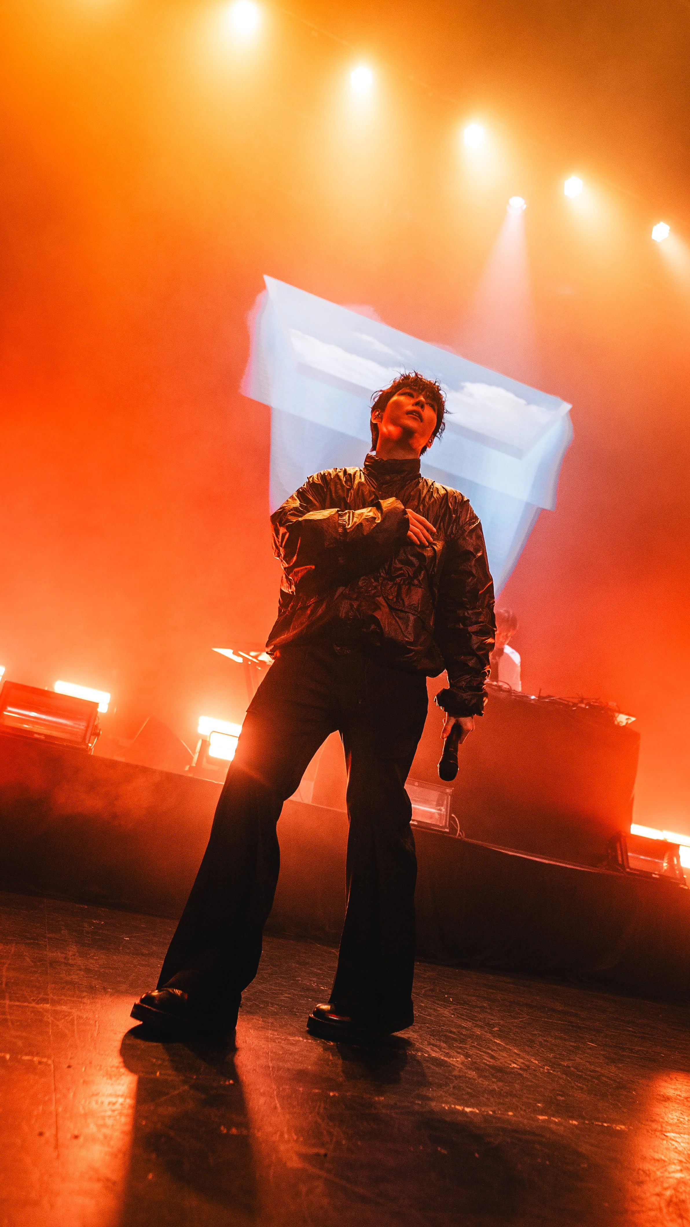 Performer on stage with orange lighting, holding microphone, wearing black outfit, with an abstract projection behind him.