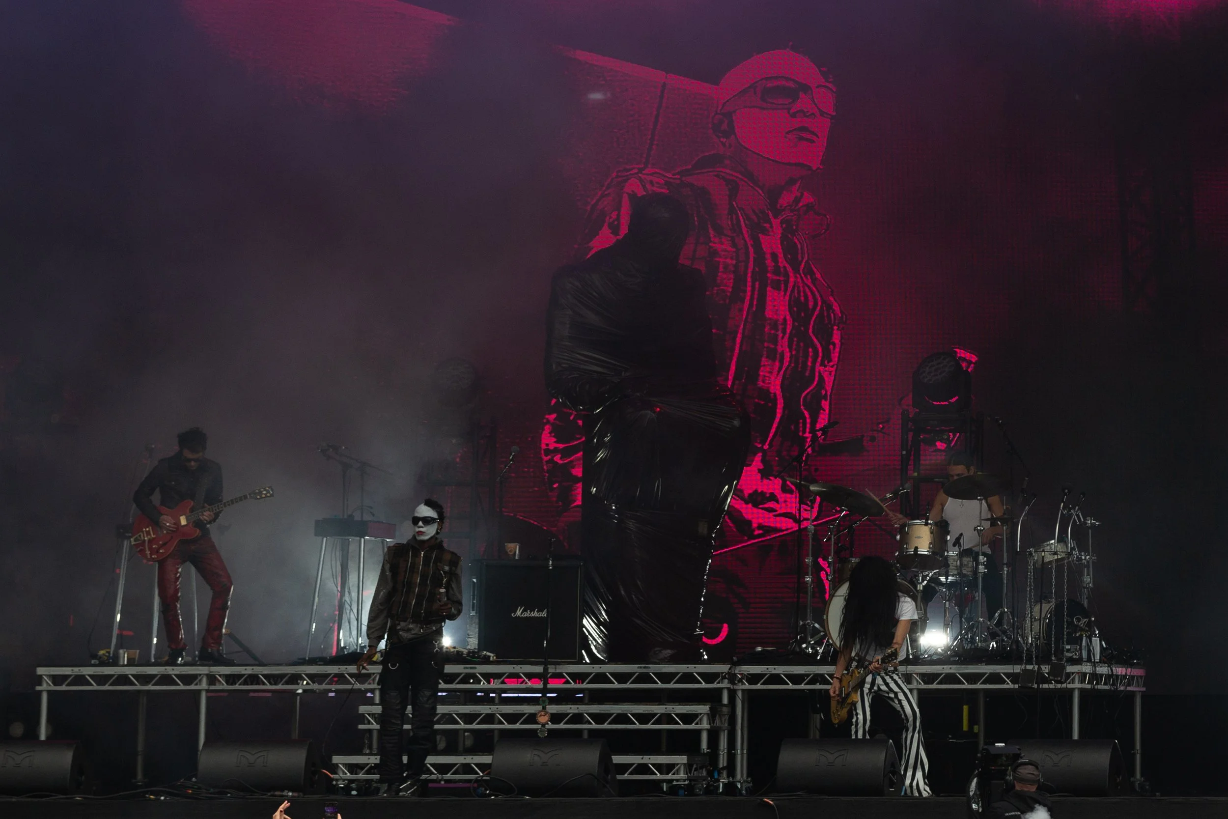 Band performing on stage with a large screen behind them displaying an image of a person wearing glasses and a jacket.