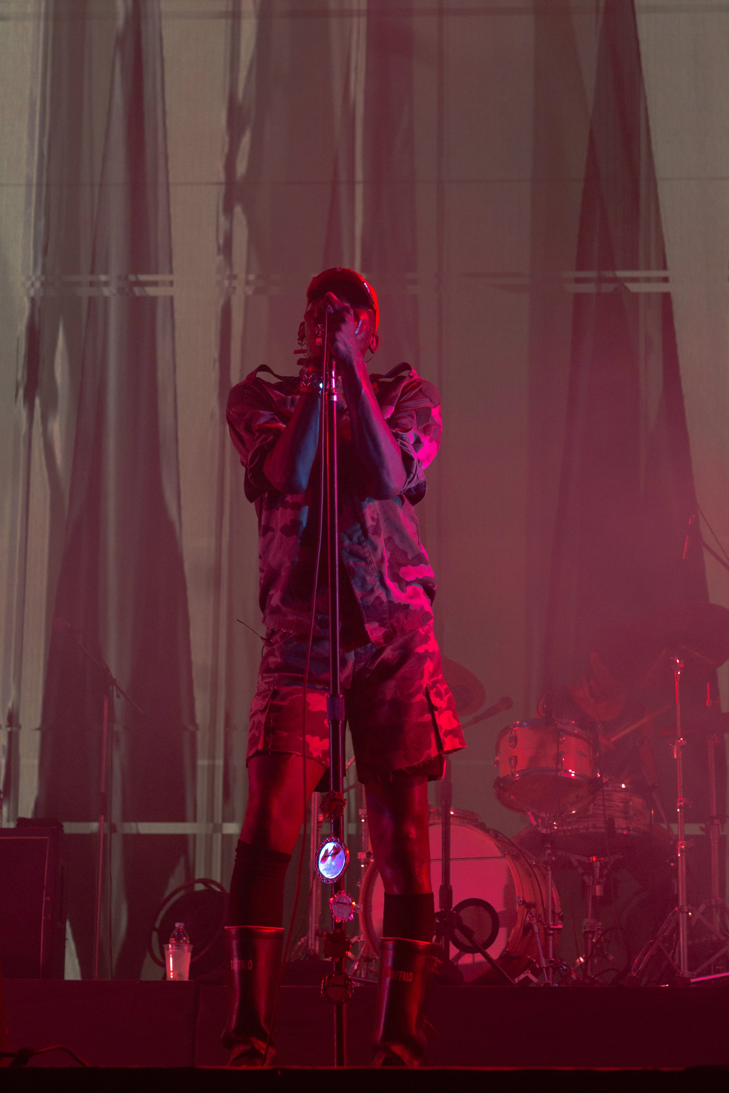 A singer wearing camouflage shorts, tall boots, and sunglasses performs on stage with a microphone, with a drummer playing in the background. The stage is lit with red and purple lights.