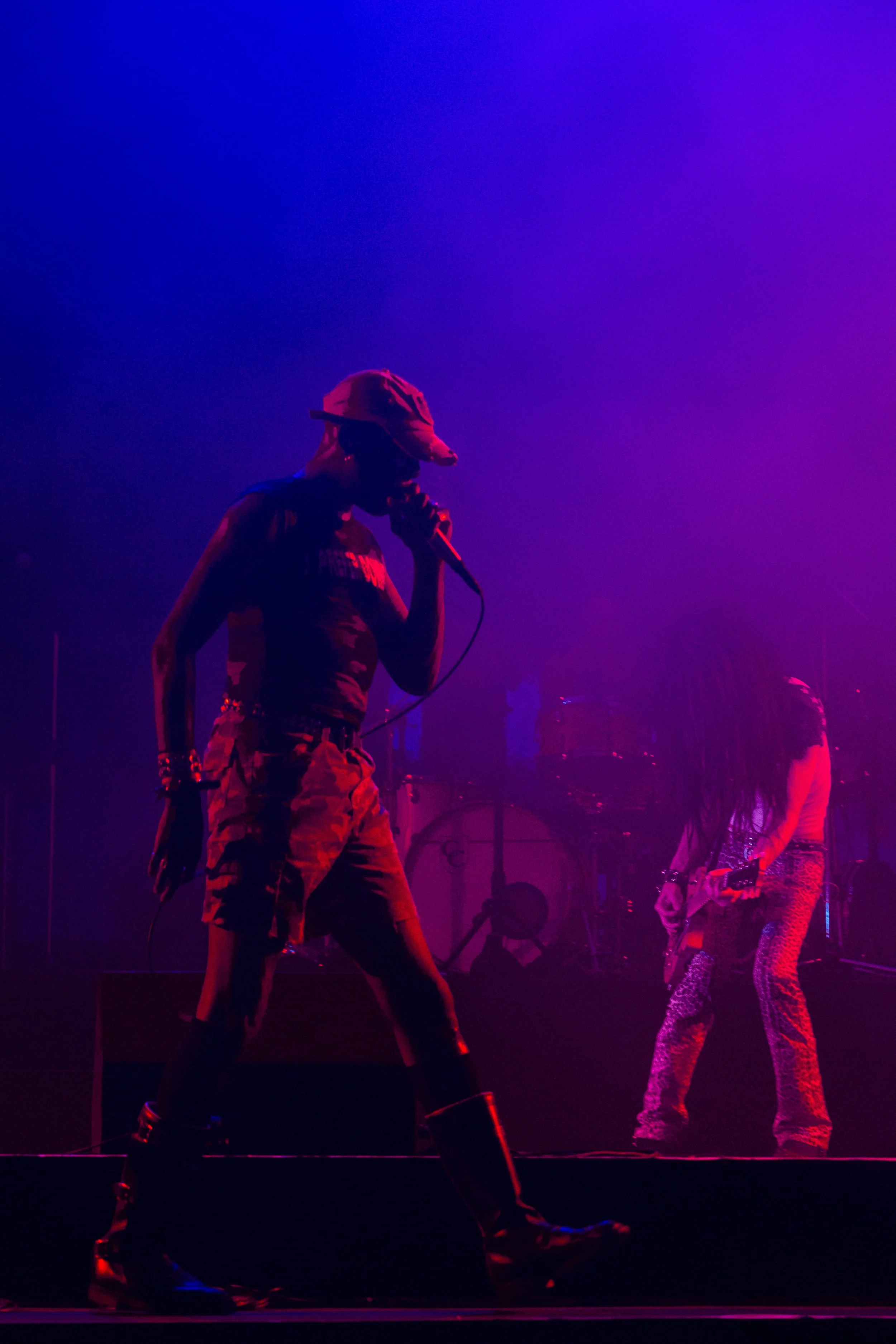 A performer singing on stage with purple and pink lighting, accompanied by a guitarist in leopard print pants.