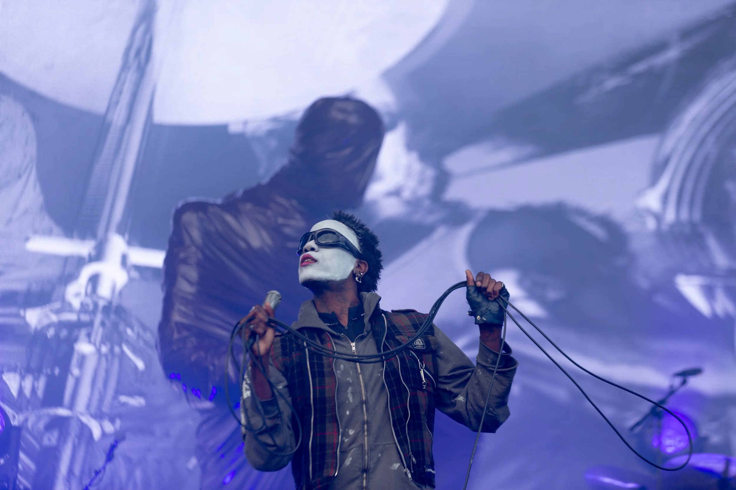 A performer with face makeup and sunglasses holding a microphone and cords on stage, with a large screen in the background projecting his image.