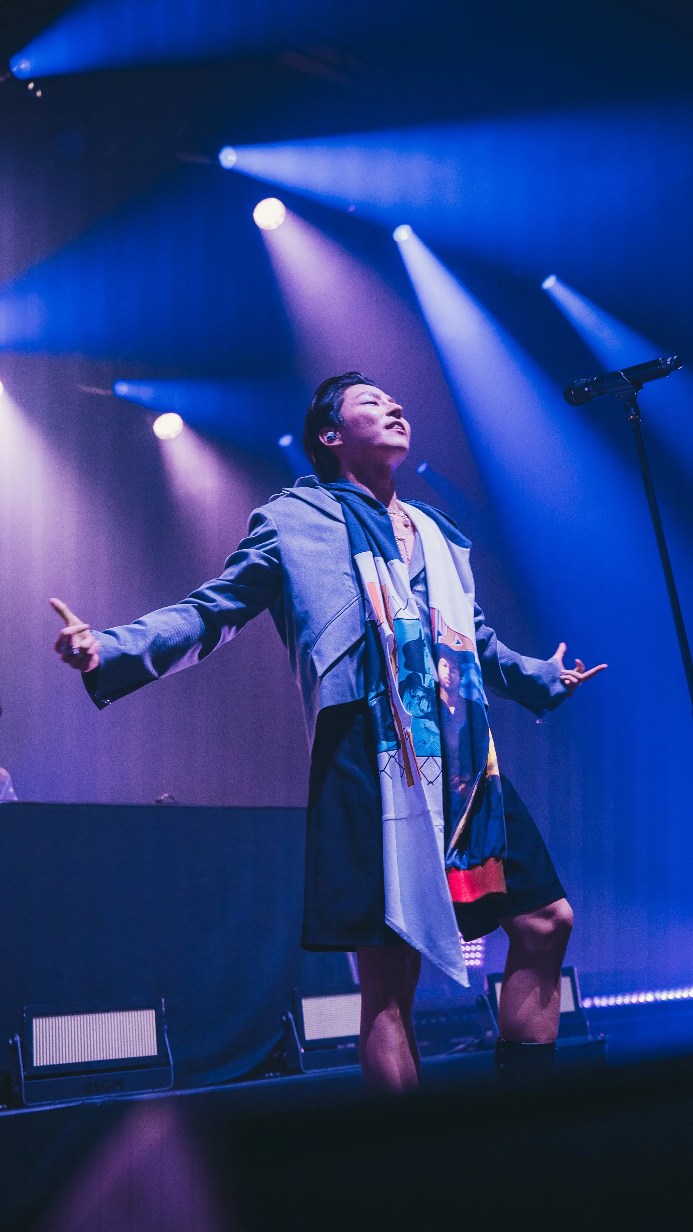 A performer on stage under colorful spotlights, wearing a jacket with a graphic tee and scarf, with arms outstretched and eyes closed, in a concert setting.