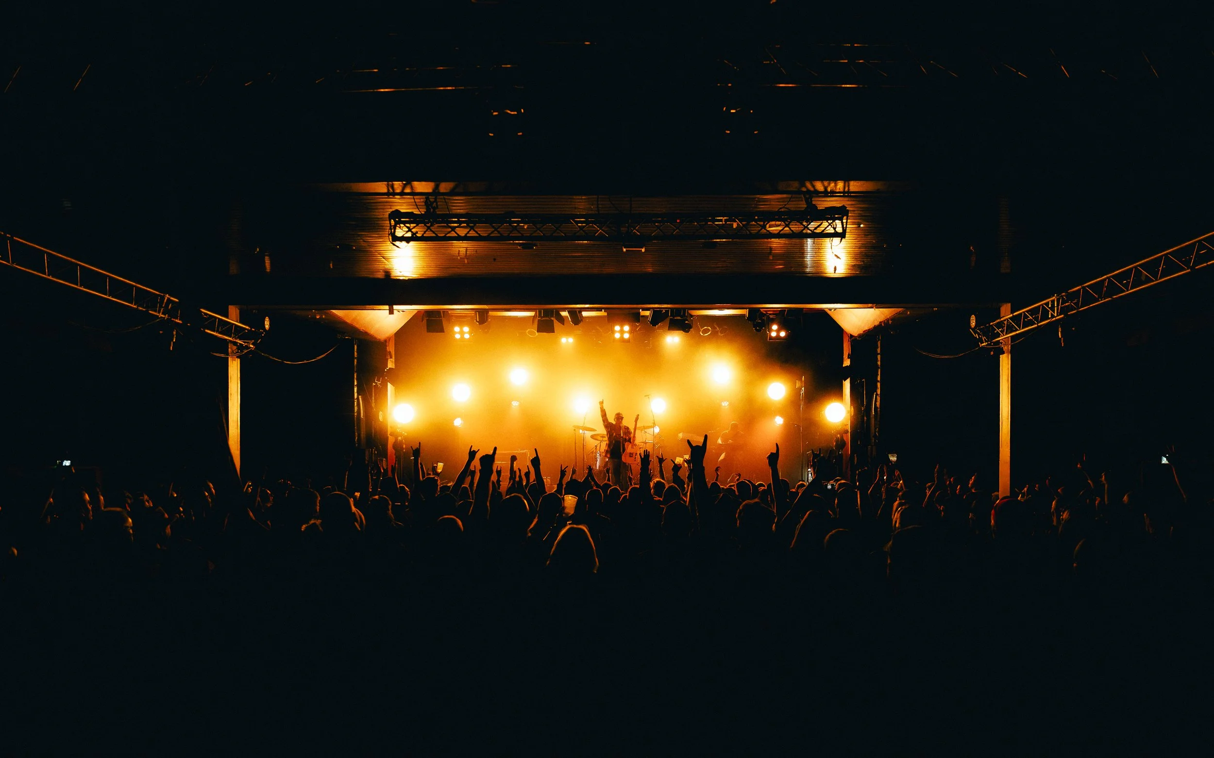 A concert with a band performing on stage illuminated by bright yellow lights, and an audience raising their hands in front of the stage.