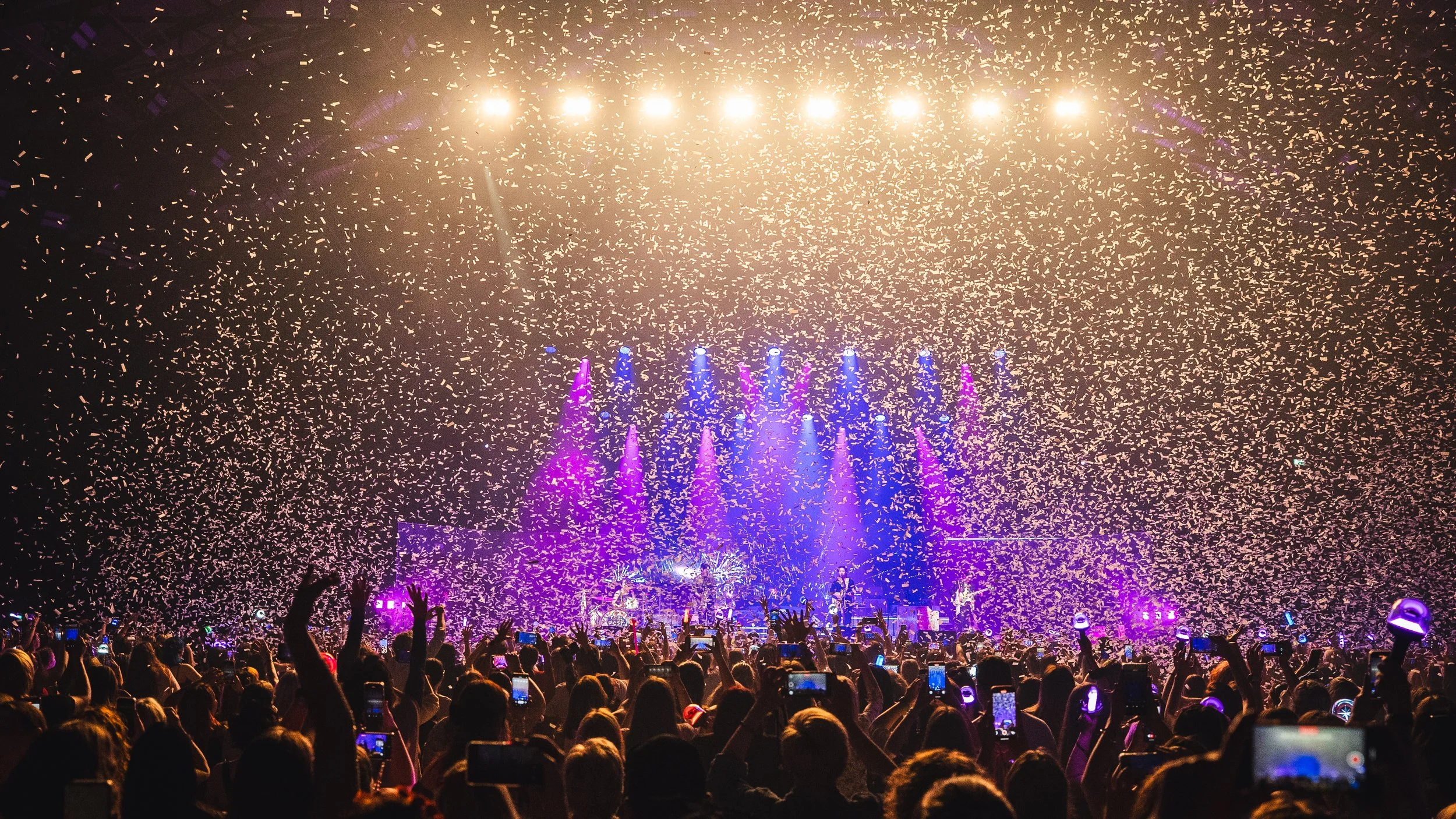 Crowd at a concert with confetti, colorful lights, and a stage in the background.