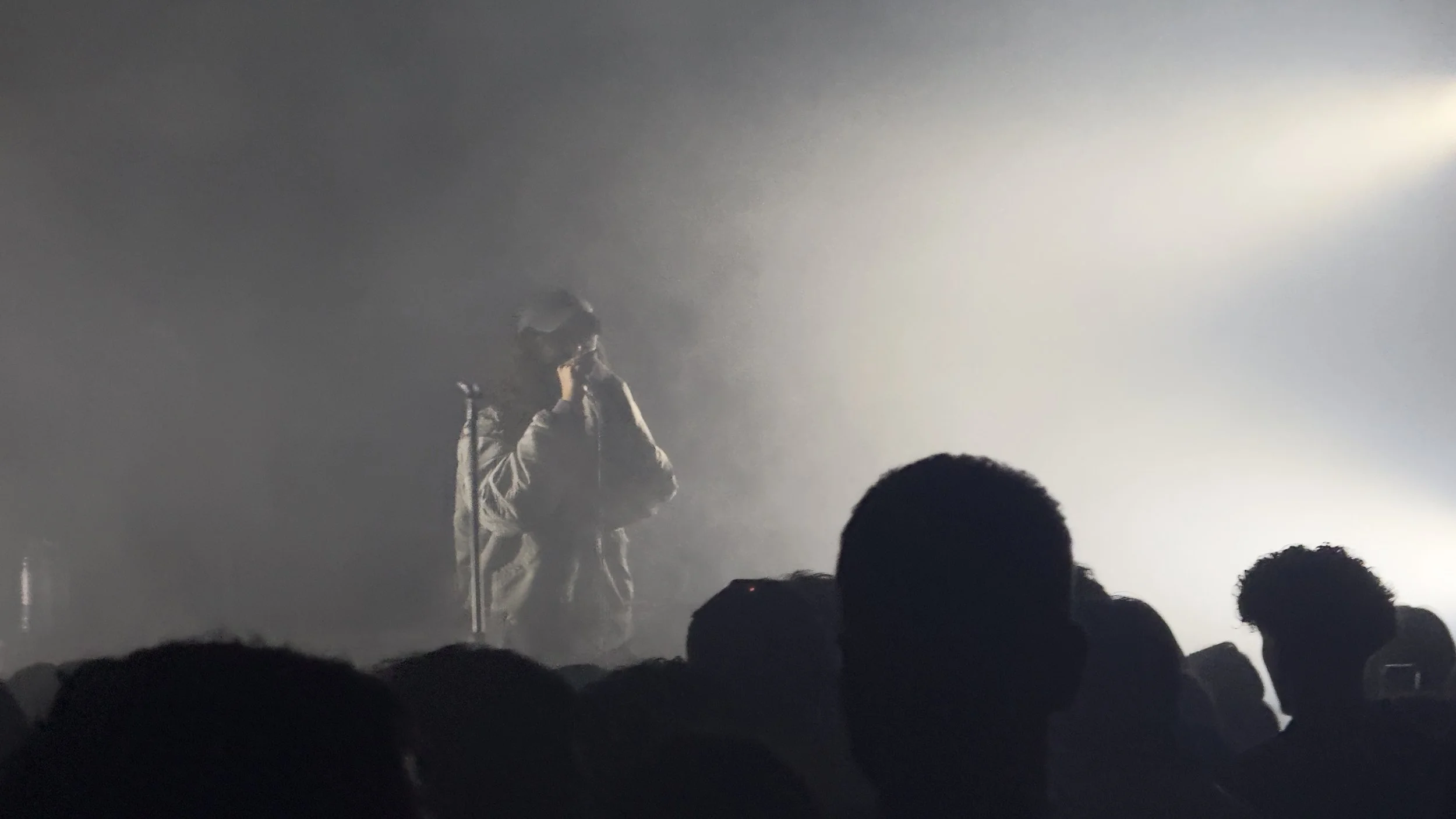 A person in a hoodie and glasses performs with a microphone on a foggy, dimly lit stage with an audience in the foreground.