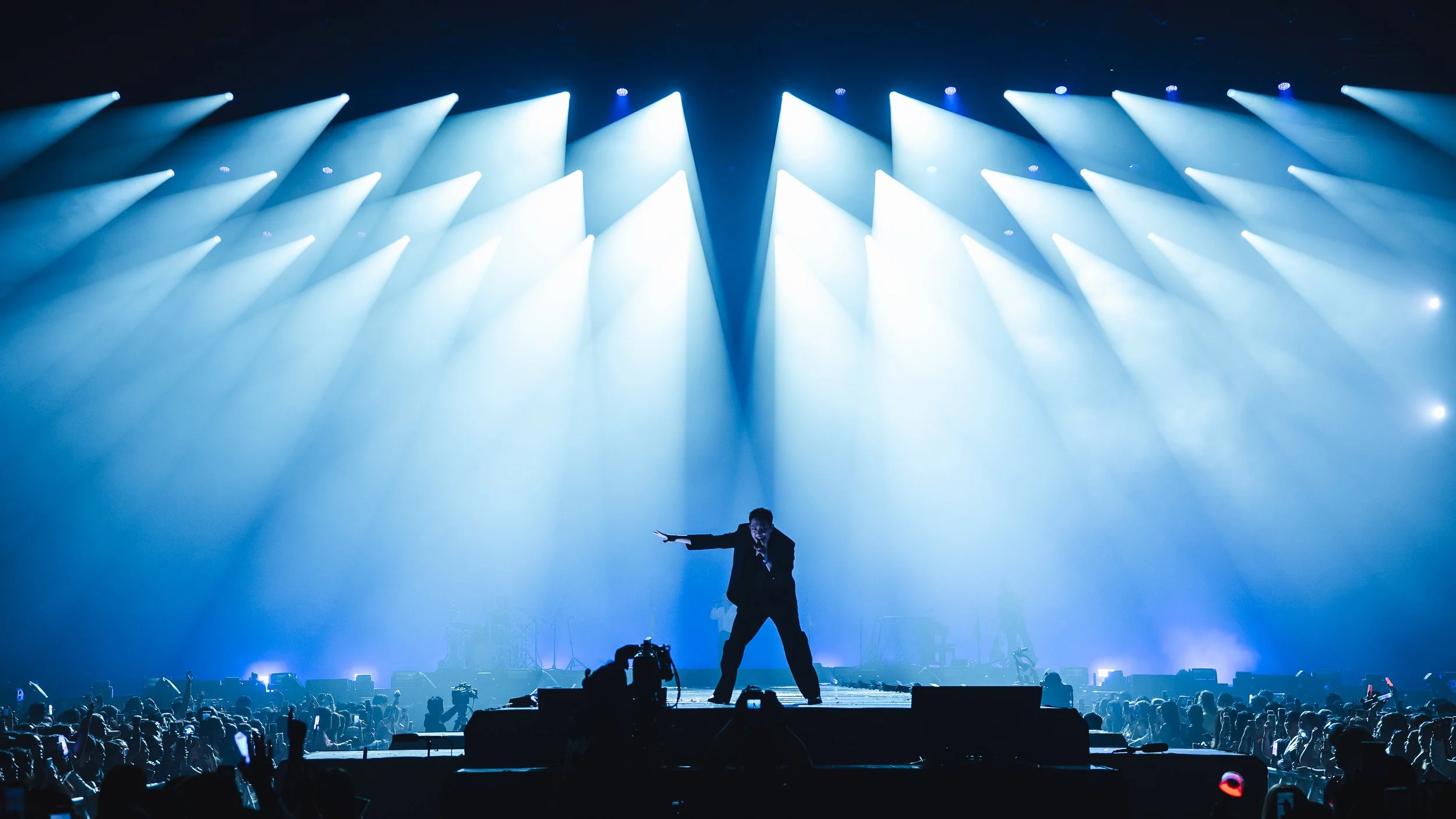 A performer standing on stage with a microphone, pointing or gesturing towards the audience, illuminated by bright blue stage lights, with a large crowd watching and recording from the front.