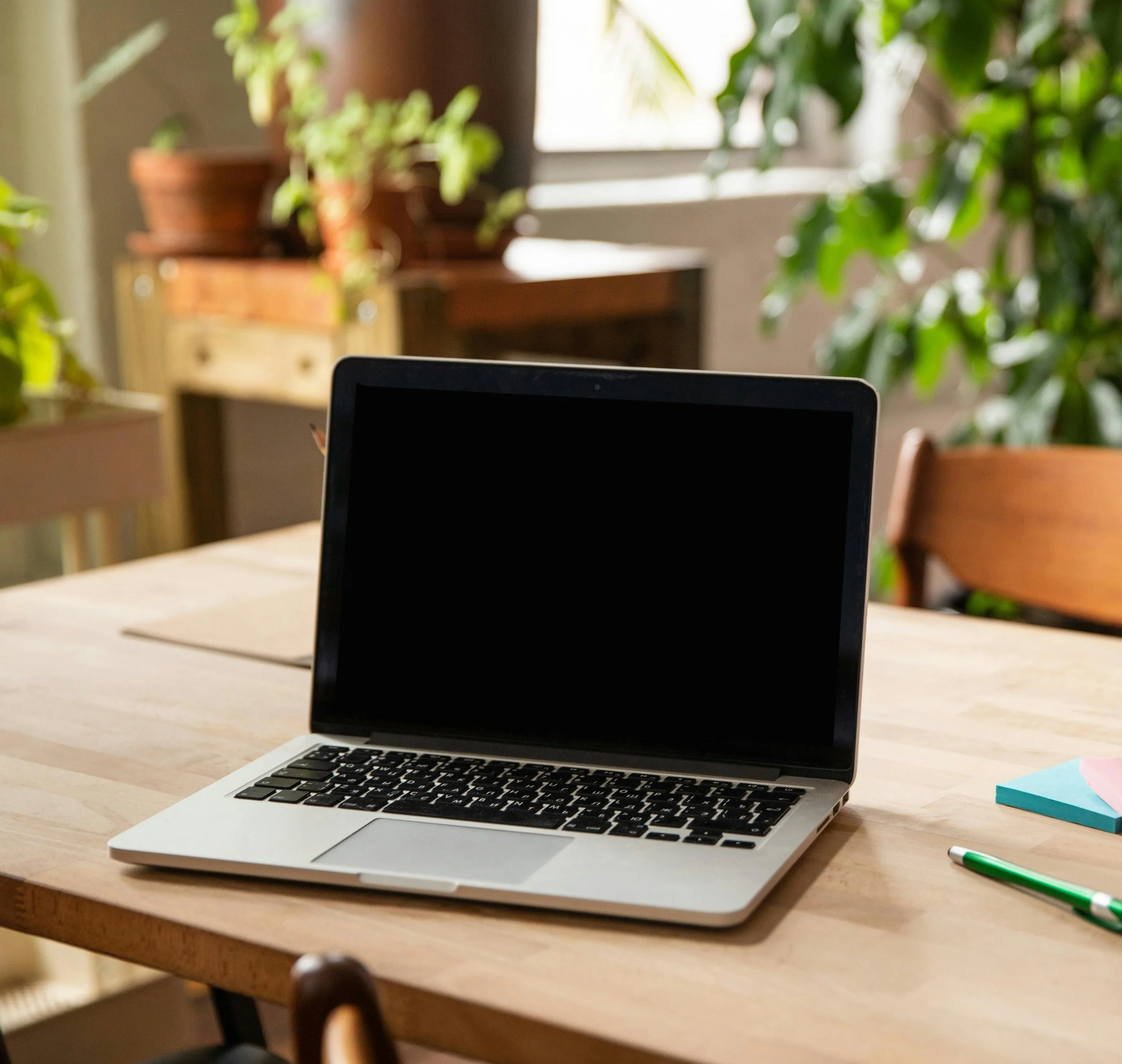 Laptop on wooden table with a blank screen, surrounded by notebooks and a pen, in a room with houseplants and natural light coming through windows.