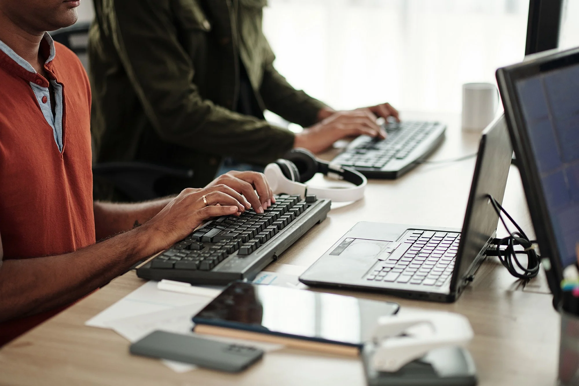 Two people working at a shared desk with laptops, keyboards, and a smartphone, in an office setting.
