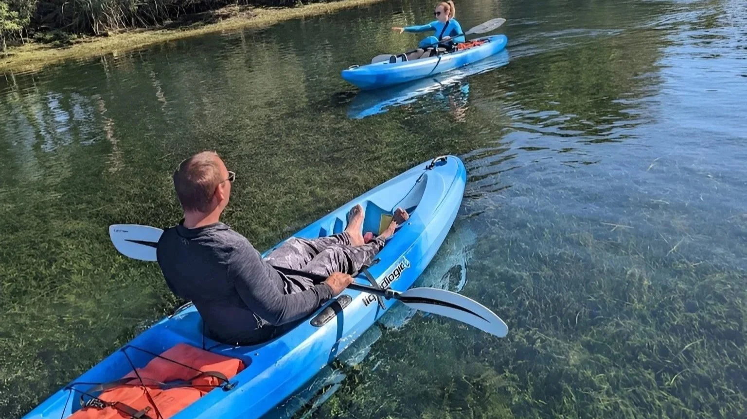 Two people in kayaks on a calm, clear lake with submerged aquatic plants visible underwater. One person is sitting and reading a book in a kayak, while the other is kayaking in the background, pointing ahead.