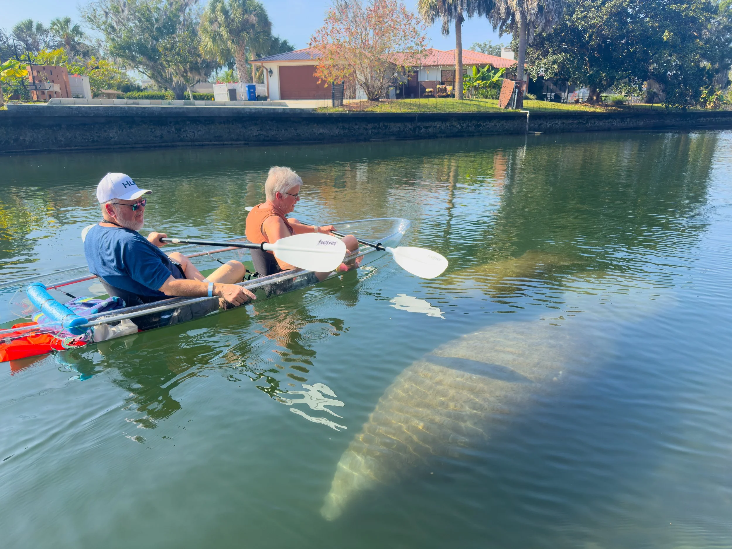 Two older adults, a man with a white beard wearing sunglasses, a white cap, and a blue shirt, and a woman with short gray hair in sunglasses and an orange tank top, are sitting in a kayak on a calm body of water. The kayak has paddles, a towel, and o