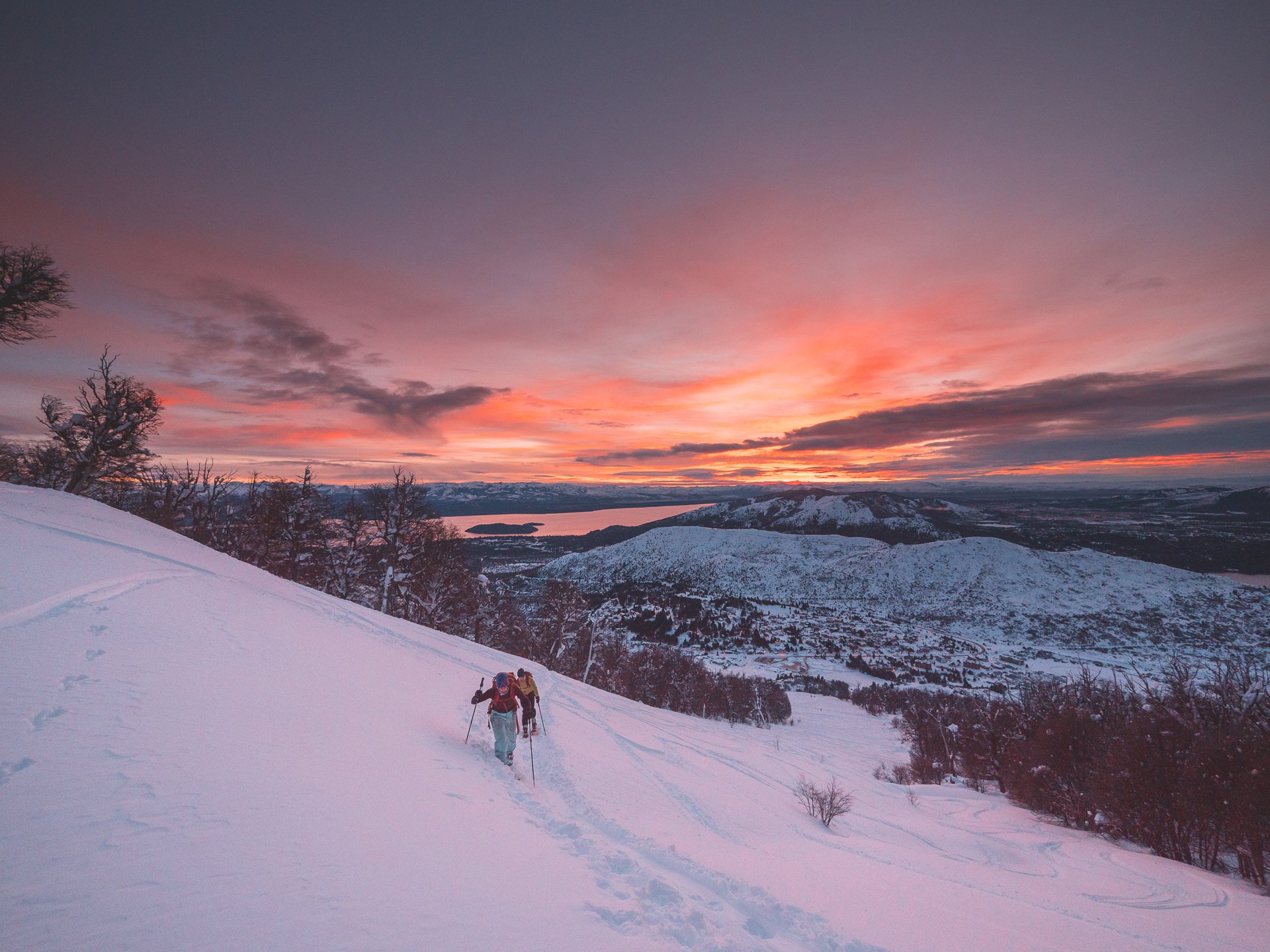 Two skiers ascending a snowy mountain slope at sunset, with a view of a valley and red-orange sky.