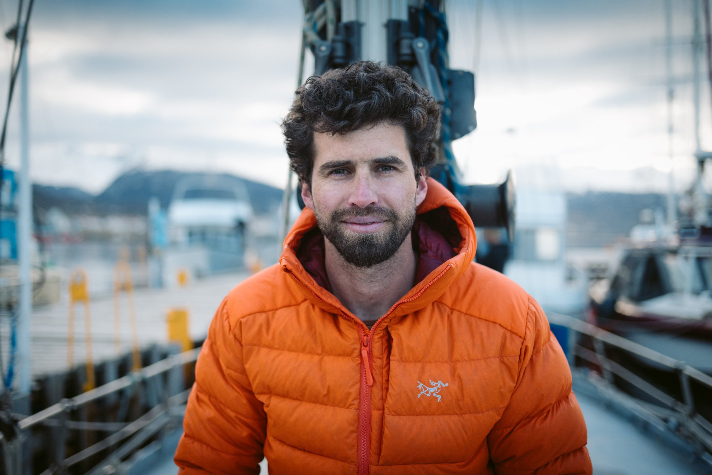 Man wearing orange jacket on a boat, with mountains and water in the background.