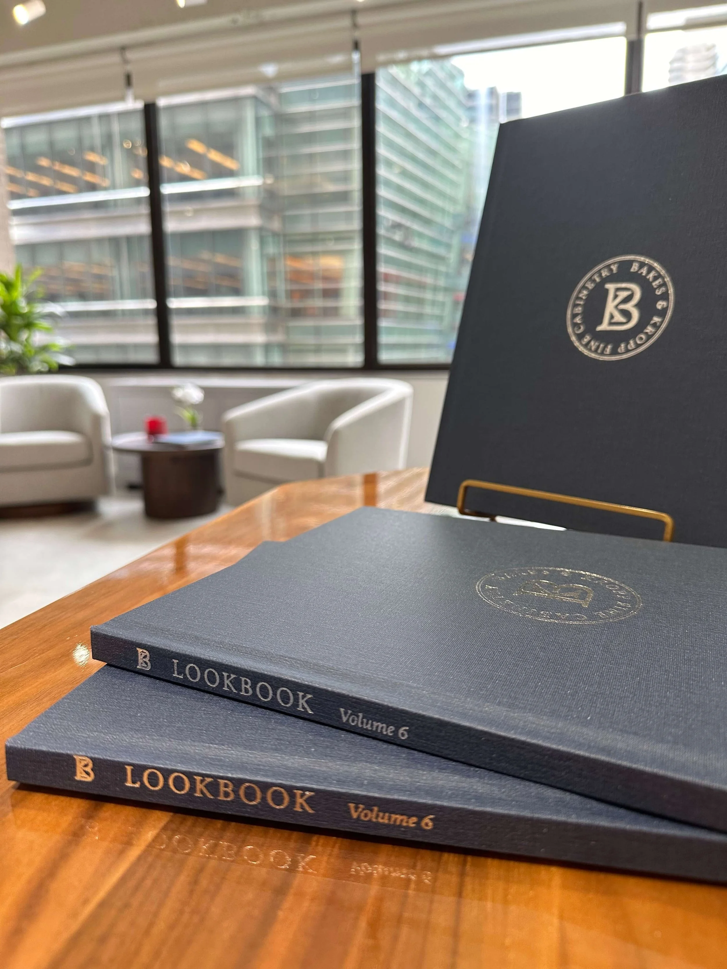 Two dark-colored hardcover books titled 'LOOKBOOK, Volume 6' placed on a wooden table, with a background of a modern office lounge featuring white armchairs, a small round table with a flower and a red object, and large windows with a view of a glass building.