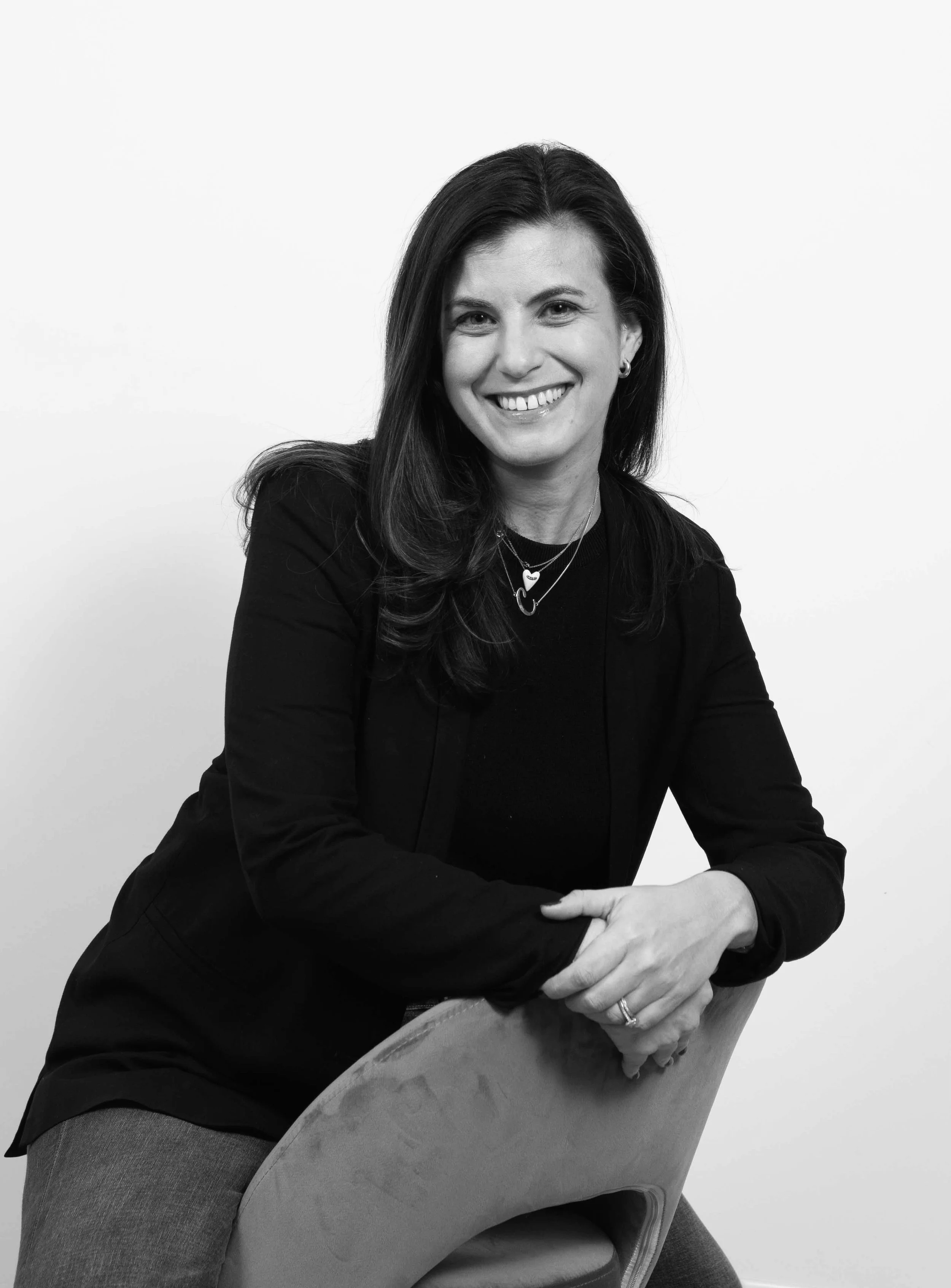 Black and white portrait of a smiling woman with dark hair, wearing a black blazer and layered necklaces, sitting on a chair with a plain white background.