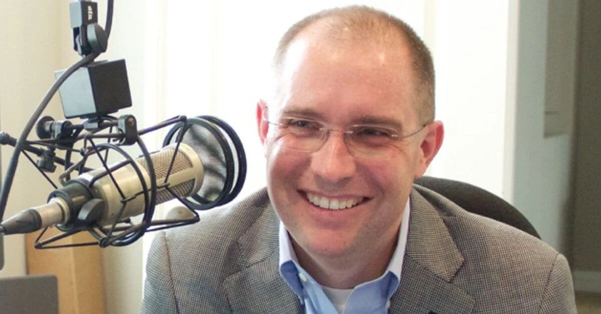 A man with short hair, glasses, and a light-colored suit jacket is smiling while sitting in front of a professional microphone in a studio or office setting.