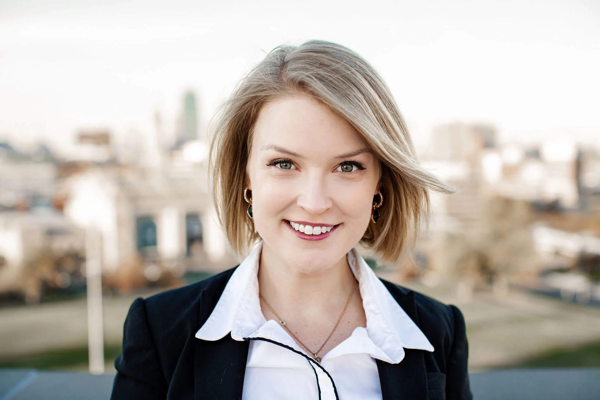 A young woman with short blonde hair and blue eyes smiling outdoors with a city skyline in the background.
