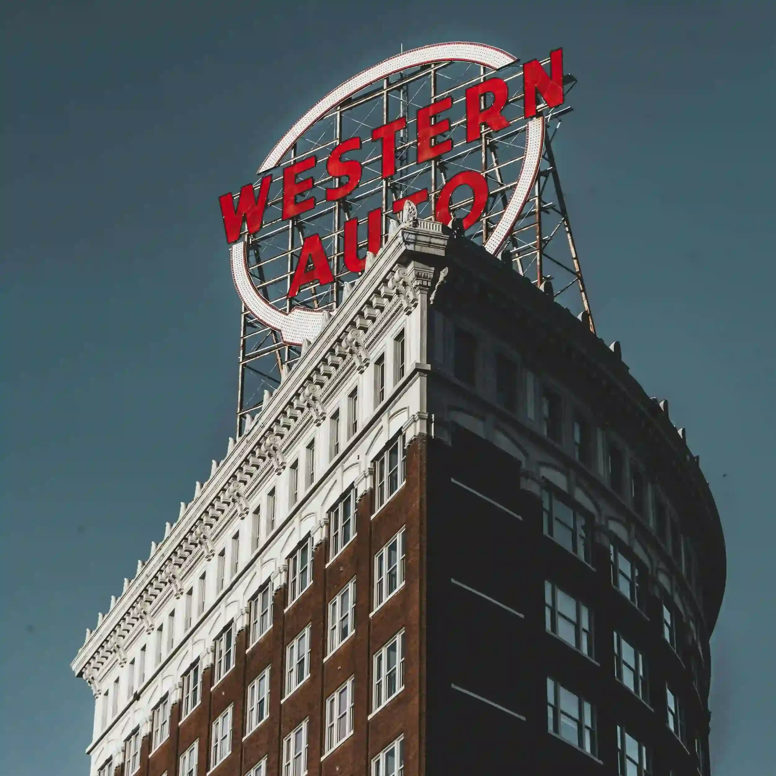 A tall building with a white and brown facade topped with a large red neon sign reading 'Western Auto' in an oval shape. The building has ornate architectural details at the top and multiple windows. The sky in the background is clear and dark blue.