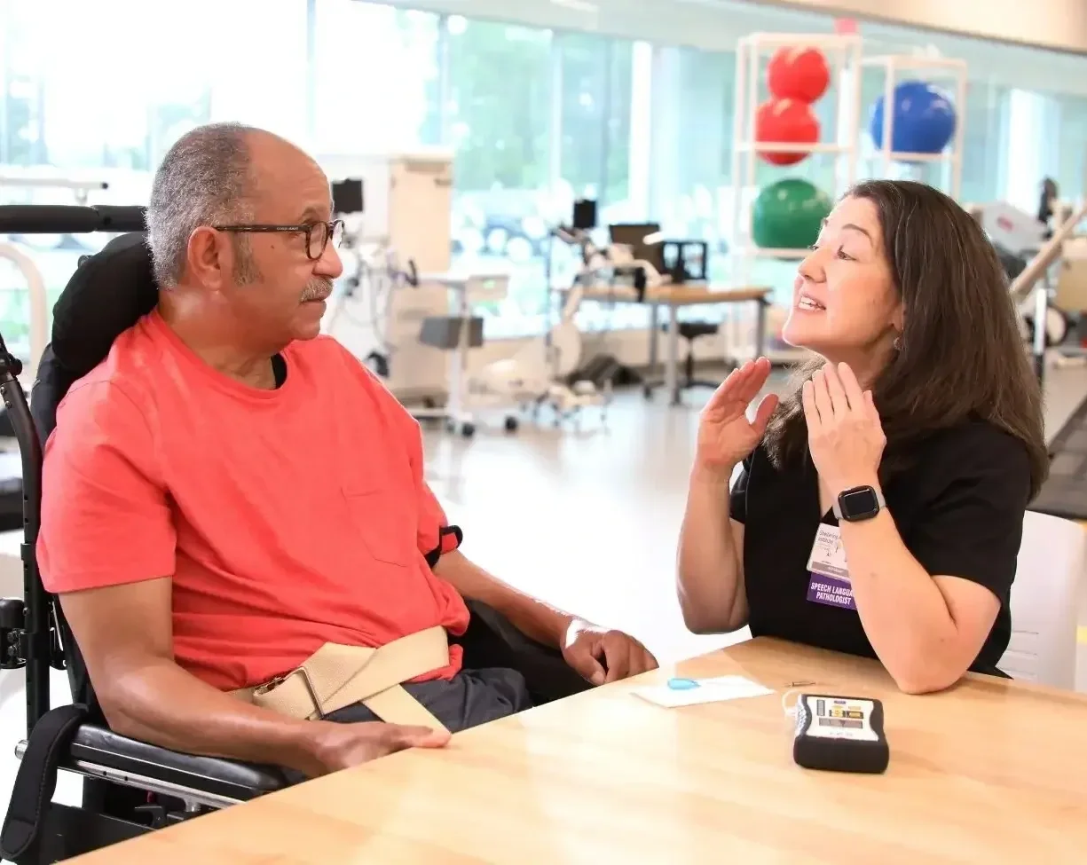 Speech-language pathologist talking to a man in a wheelchair in a therapy room with exercise balls and equipment in the background.