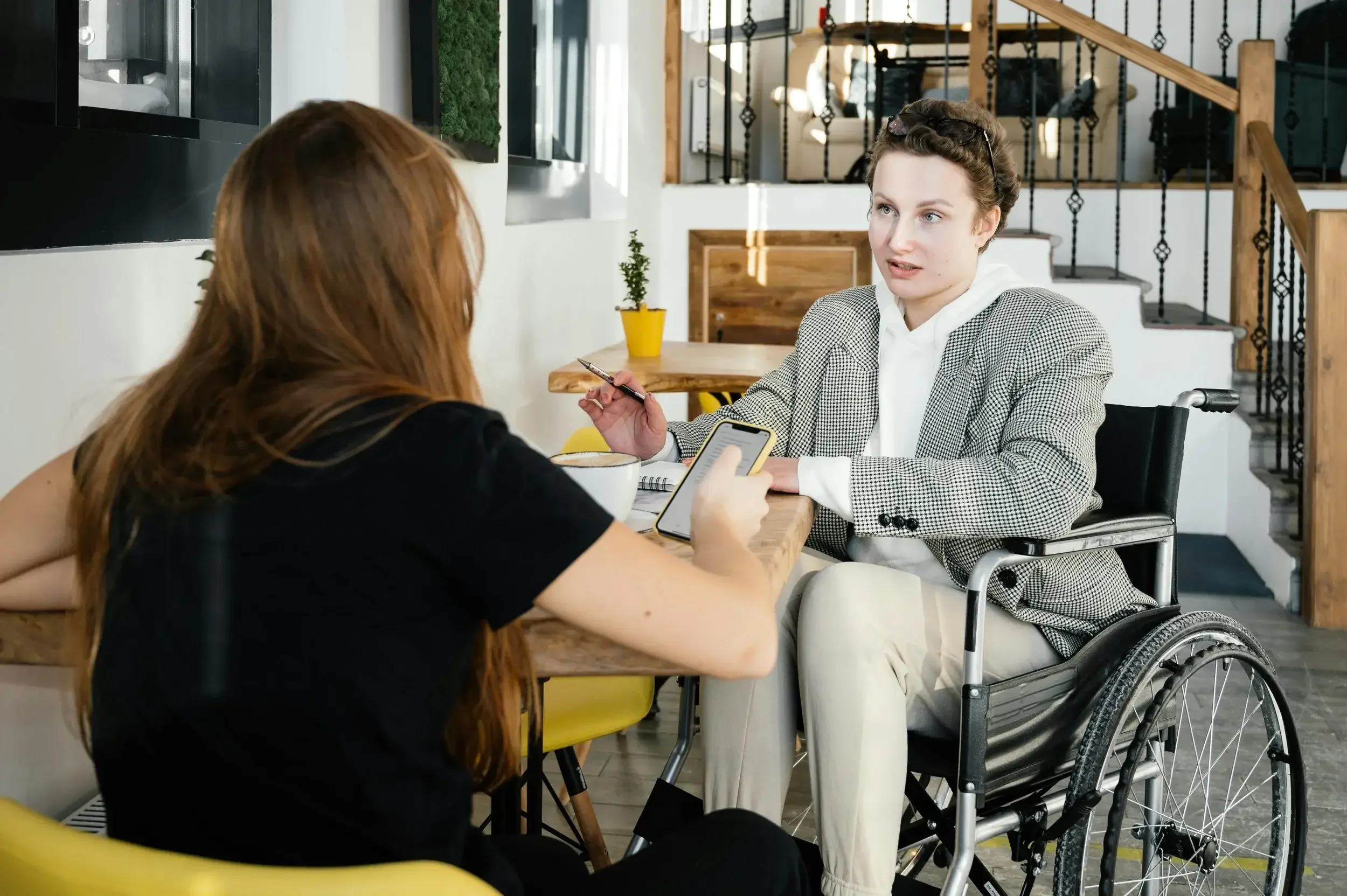 Two women, one in a wheelchair, discussing ideas at a cafe table, demonstrating inclusive communication coaching, active listening, and networking skills.