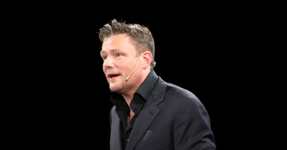 A man in a dark suit and black shirt speaking, wearing a headset microphone, with a black background.