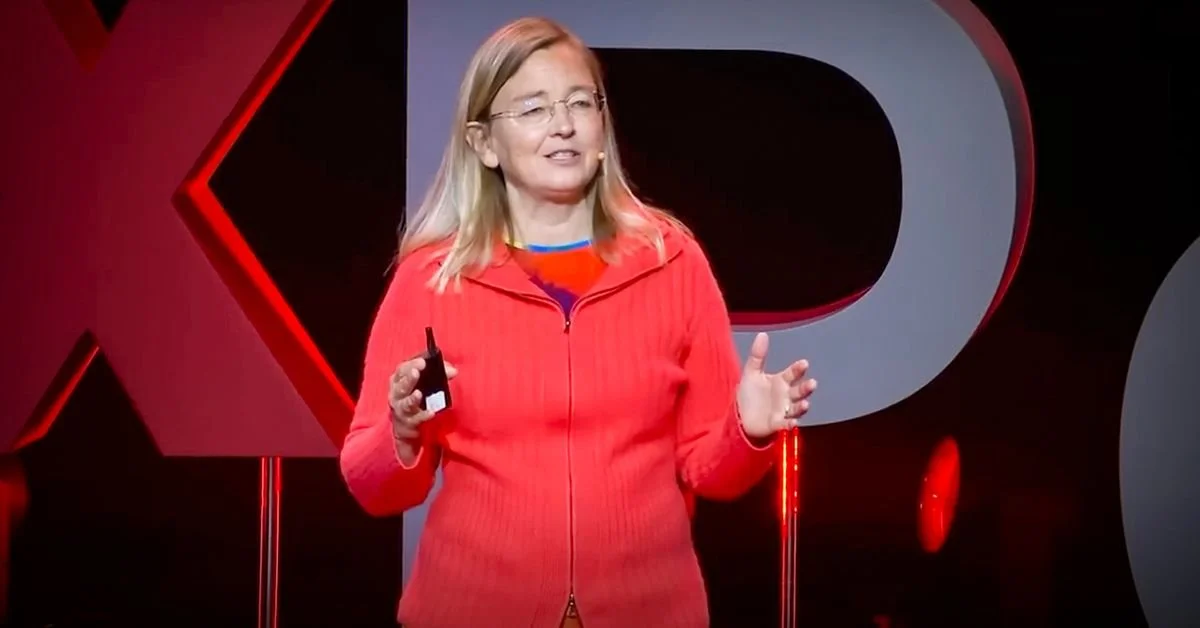 A woman with blonde hair wearing glasses and a red zip-up sweater giving a presentation on stage with a TEDx logo in the background.